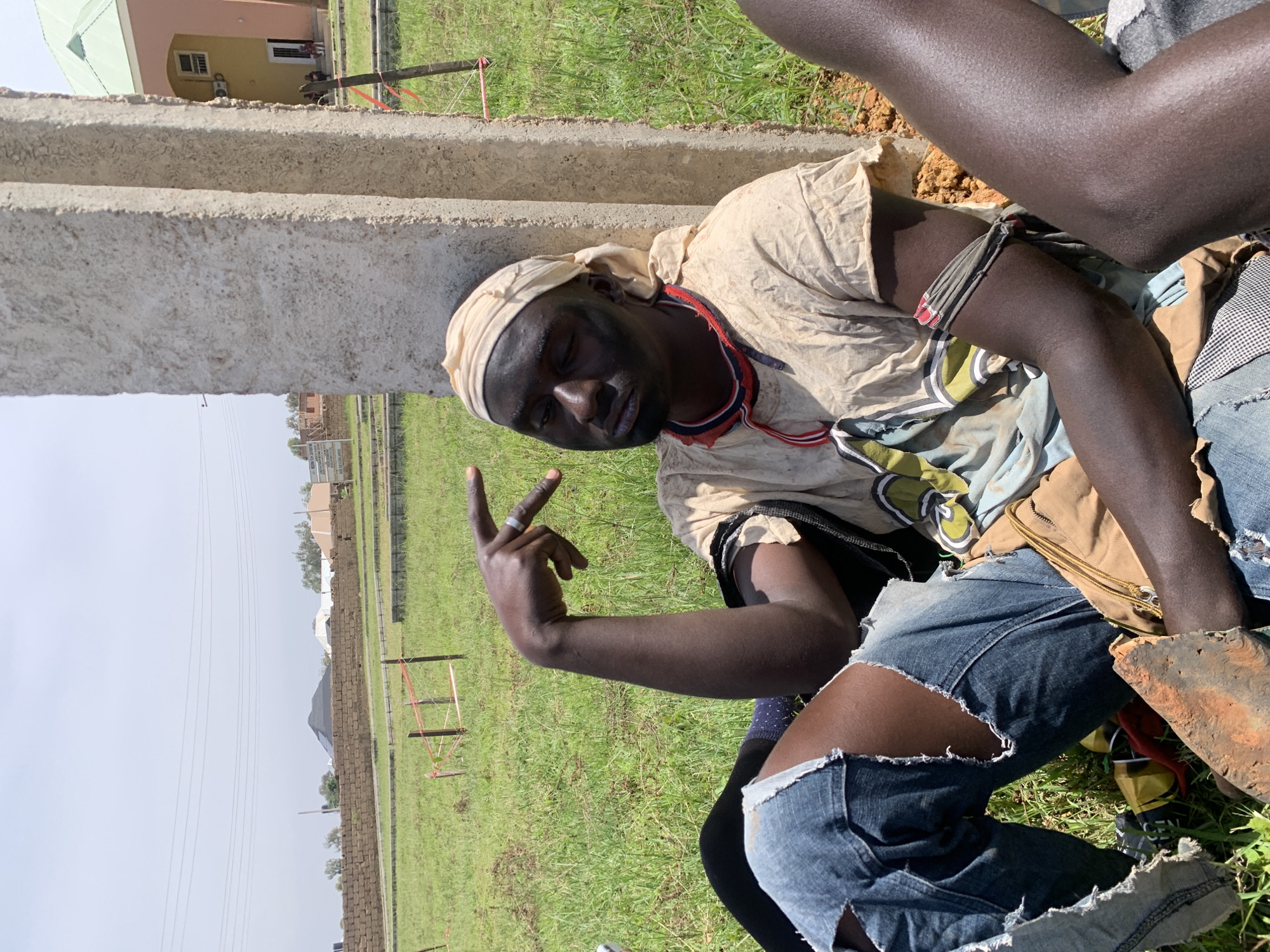 In Gwafan, Nigeria, a dark-skinned individual leans against a concrete pillar, their head tilted back with eyes closed. They wear a tan-colored shirt with a patterned trim around the neck and a light-colored headscarf. Black face paint is visible on their face. Ripped blue jeans are worn, and an arm is extended with fingers flexed, adorned with a ring. Another person's leg is visible to the right, showing dark skin. The individuals are seated on a grassy area with some sparse vegetation. In the background, there is a brick wall, construction materials, and several buildings, including one with a light pink facade and green roof. The sky is overcast.