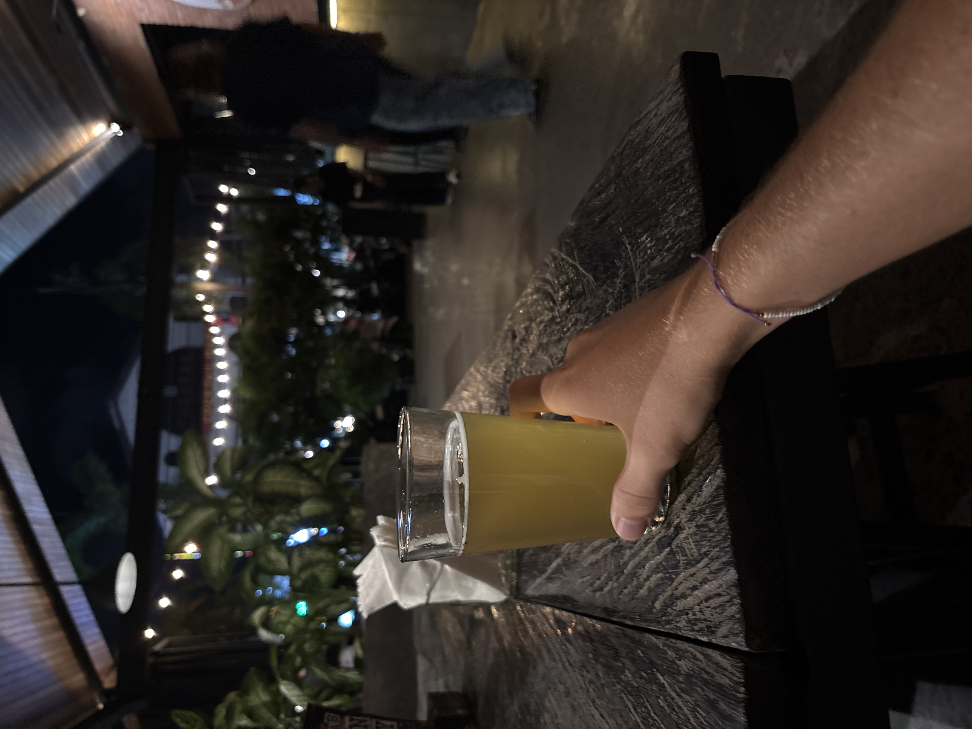 A close-up indoor shot captures a hand holding a glass of light-colored beer on a weathered wooden table. A bracelet adorns the wrist. The backdrop features string lights, and a silhouette of a person can be seen in the background. The setting is in North Kuta, Indonesia.