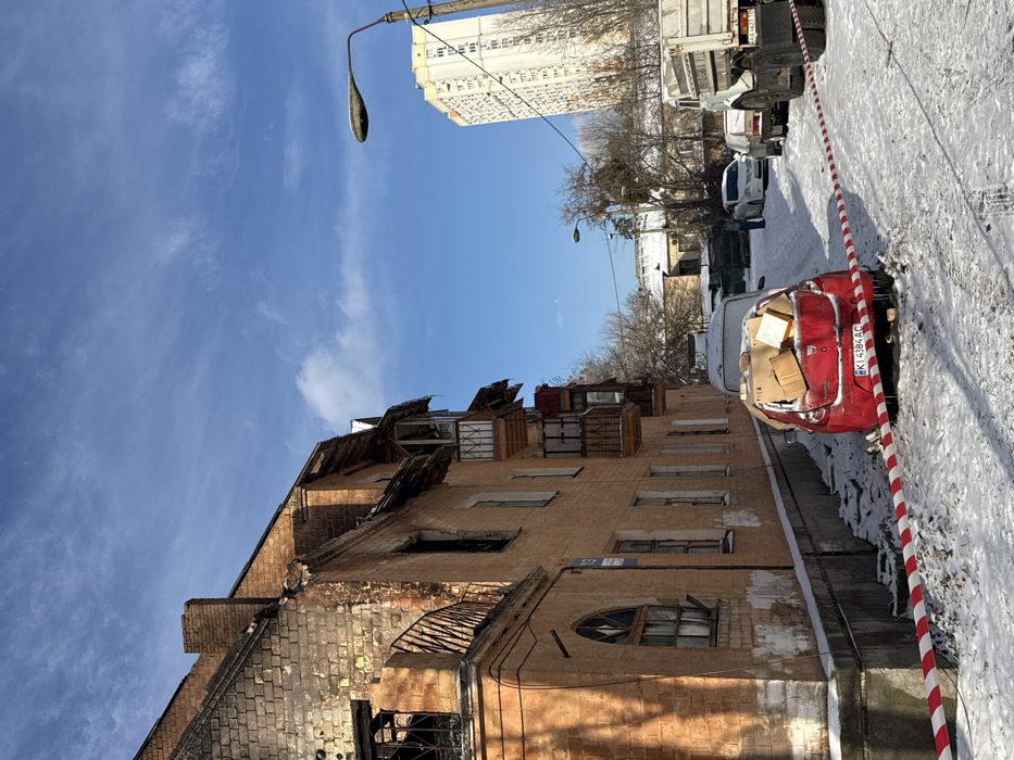 The scene depicts a snow-covered street in front of a heavily damaged residential building at Poliska Street (вул. Поліська), 13, in Kyiv, Ukraine, under a clear blue sky with wispy clouds. The multi-story brick building on the left exhibits significant charring and structural damage, particularly on its upper floors and roof, consistent with fire. Blackened brickwork is visible, and several windows are broken, some with missing frames. Icicles hang from damaged balconies and roof edges.
In the foreground, a red and white striped barrier tape marks off a snowy area. Behind the tape, a red Dacia Sandero hatchback (Ukrainian license plate KI 4384 AC) is parked, partially covered in snow. Its rear window is absent, and the interior is visibly filled with several cardboard boxes. Further down the street, also covered in snow, are a white sedan, another darker sedan, and a large, light-colored dump truck on the right edge of the frame. One individual, dressed in dark clothing, stands near the white sedan in the middle ground. A tall, light-colored multi-story building is visible in the background, to the right of a street light pole. Bare trees and smaller, snow-covered structures complete the background. The ground is consistently covered in snow, showing tire tracks and footprints.