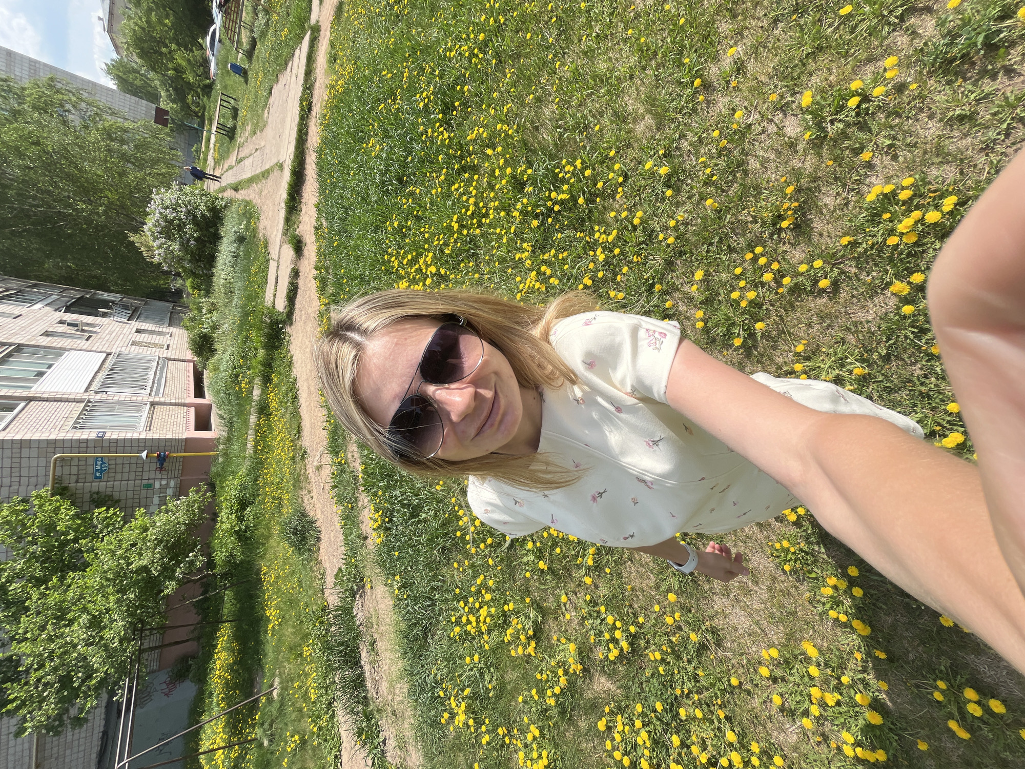 A woman takes a selfie outdoors in Chaikovsky, Russia. She is in a field of dandelions wearing sunglasses and a light-colored dress. A residential building is visible in the background. A walkway leads through the field, and trees are also present.