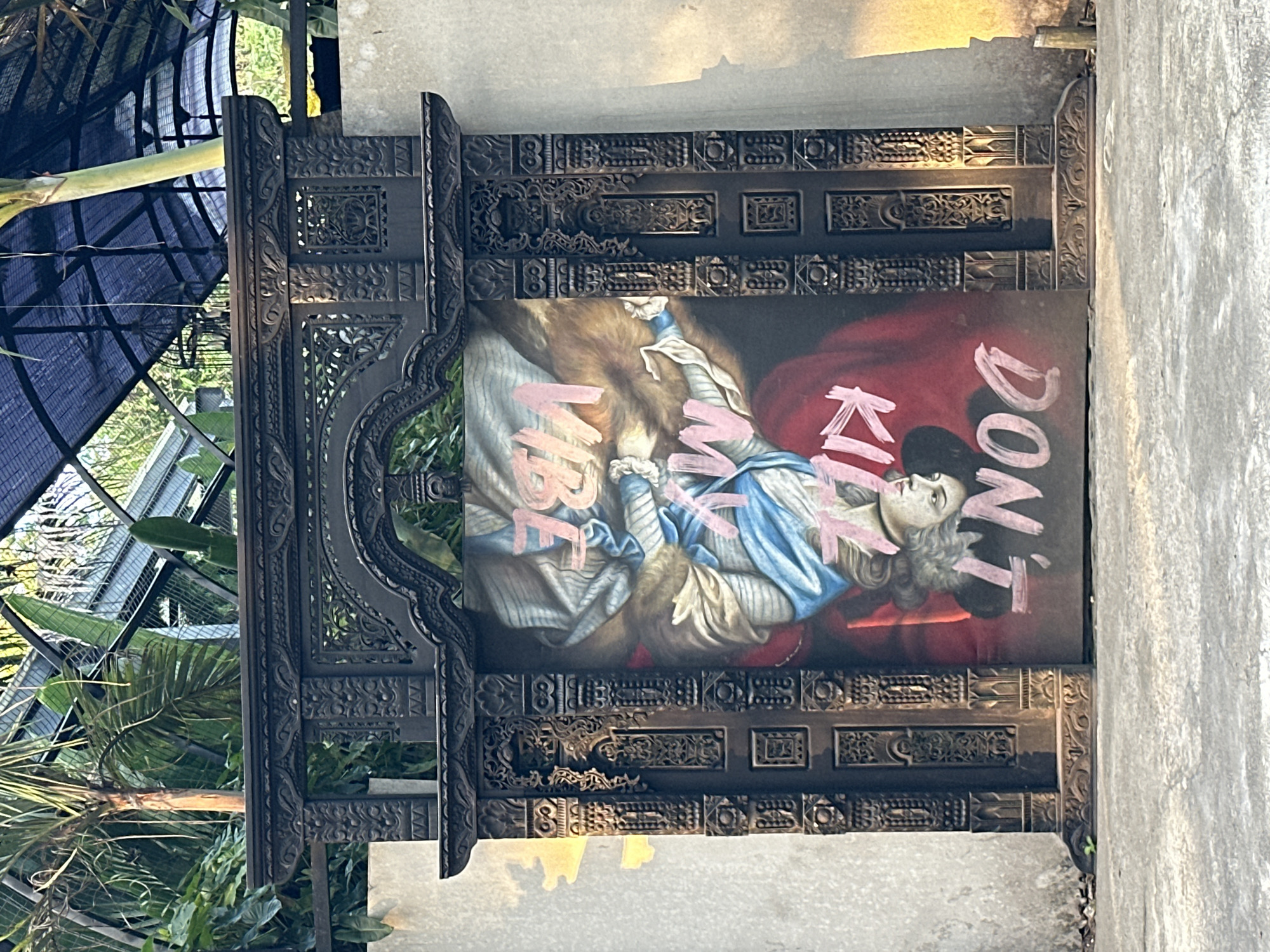 An ornate, dark wooden archway stands in front of a concrete wall. Inside the archway is a painting of a figure that appears to be upside down, with pink text overlayed on the painting. The text reads "VIBE MY KILLA DON'T". The painting depicts a person with long blonde hair wearing a blue and white garment and a red cloak. They are looking downwards. The archway is intricately carved with floral and geometric patterns. Behind the archway, lush green foliage and palm trees are visible, suggesting an outdoor, possibly tropical, setting. The ground in front of the archway is paved with concrete. Sunlight casts shadows on the wall to the right of the archway. The scene appears to be located in Kuta Selatan, Indonesia, possibly at a tourist attraction or a cultural site.