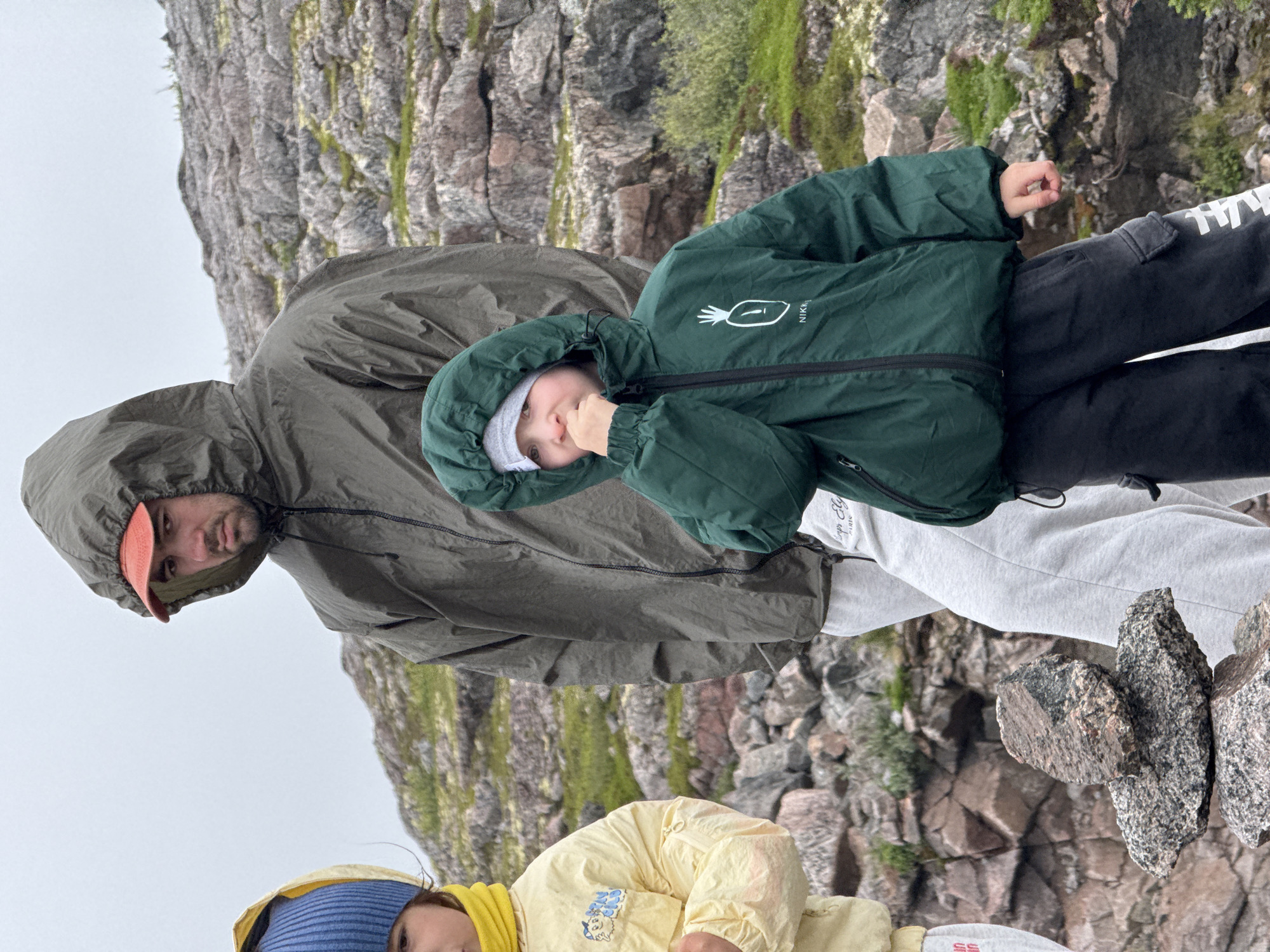 In Teriberka, Russia, three individuals stand amidst a rocky, moss-covered landscape under an overcast sky. In the foreground, a child wearing a dark green hooded jacket with a white pineapple graphic and the brand name "NIKK" visible, is looking towards the camera with their hand near their mouth. To the left, another child in a light yellow hooded jacket and a blue beanie is partially visible. Behind them, an adult man, wearing a dark olive green hooded jacket and an orange cap, faces away from the camera, looking down. The ground is covered in scattered rocks, and a cairn of stacked stones sits prominently in the lower left of the frame.