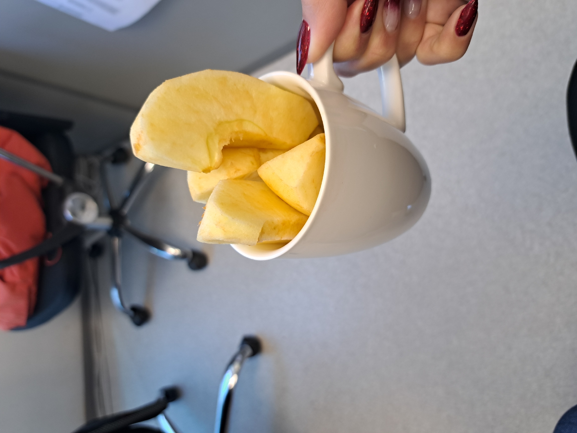 A hand with long, red, glittery fingernails holds a white mug filled with cut pieces of a light yellow fruit, likely apple or pear. The background shows a light gray wall and part of an office chair with a red cushion. The image appears to be taken indoors. The location in Sofia, Bulgaria cannot be determined from the given visual information.