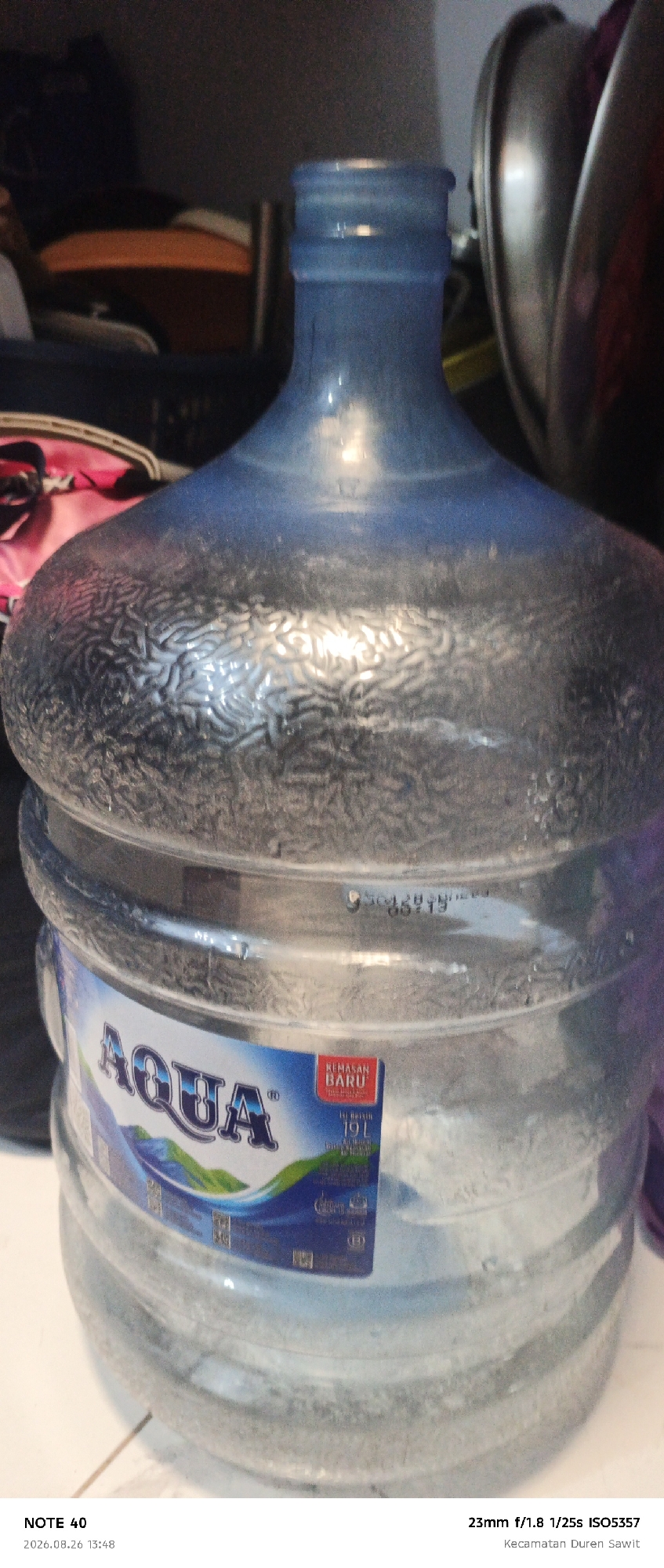 A large, blue-tinted plastic water jug is the primary subject of the image. The jug is full of water and has a textured pattern on its upper half. A label on the lower half of the jug reads "AQUA" in large, stylized letters and also shows the text "KEMASAN BARU" and "19 L". The jug is positioned on a light-colored tiled surface. In the background, there are blurry objects, including what appears to be a metal basin or pot to the right and some dark fabric items to the left, suggesting an indoor setting. The lighting appears to be artificial. The bottom of the image contains text indicating the device used ("NOTE 40"), the date and time of capture ("2026.08.26 13:48"), and camera settings ("23mm f/1.8 1/25s ISO5357"). Below this, the location is identified as "Kecamatan Duren Sawit", which is a district within the city of East Jakarta, Indonesia.