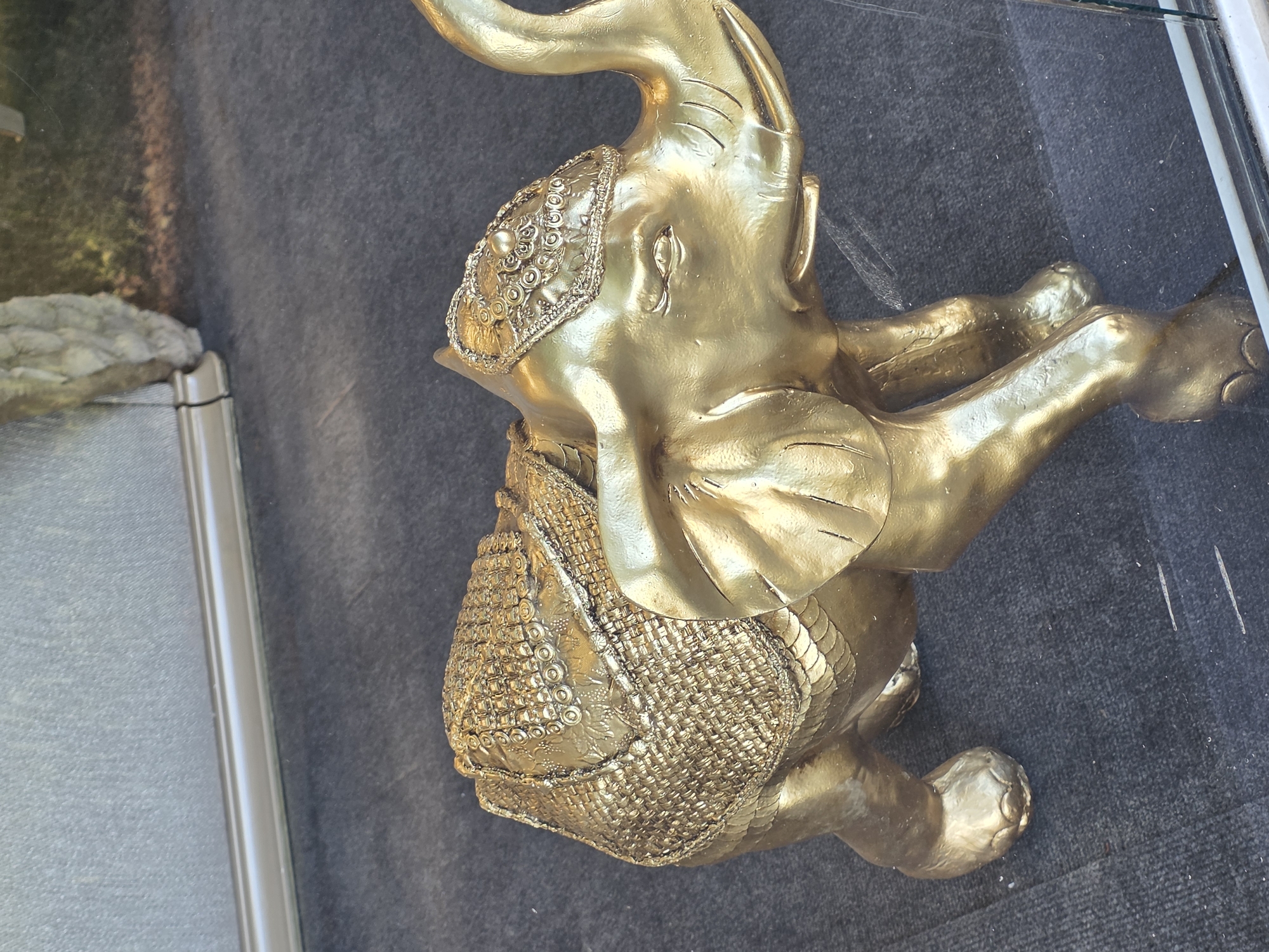 The image features a gilded elephant figurine, presented in a static, resting position with its trunk raised. The figurine is made of a shiny, metallic gold material, reflecting ambient light. Intricate decorative patterns adorn the elephant's head and body; the head features embossed circular motifs and swirls, while the body is covered with a textured, scale-like or woven pattern that forms a ceremonial saddle.
The elephant rests on a dark, possibly carpeted or fabric surface, which shows minor specks of dust or lint. The entire scene is viewed through a glass barrier, evident from numerous reflections superimposed on the background. These reflections include glimpses of an outdoor environment, with green foliage or trees partially visible in the upper left, and a vertical metallic frame or barrier on the right side of the image. A grey, rock-like object is also reflected on the left.
The lighting suggests it is daytime, contributing to the bright sheen of the gold elephant and the clarity of the reflections. Some minor wear or small cracks are visible on the elephant's gold surface. There are no people present, and no specific activity is taking place. No visible text can be identified in the image. The location context for this image is Plovdiv, Bulgaria.