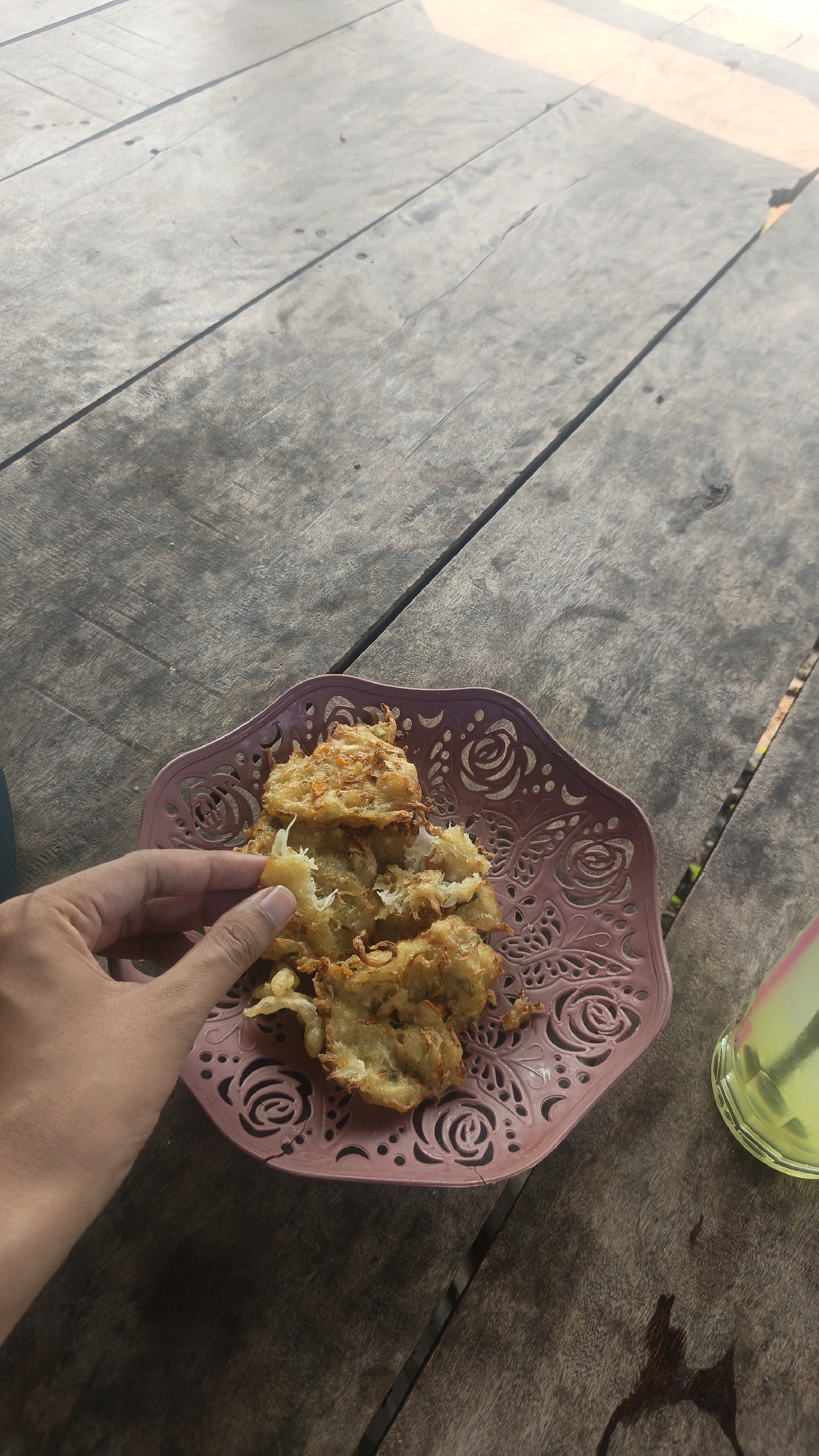 An overhead shot shows a hand holding a piece of food over a pink plate filled with more of the same food, on a wooden table. The food appears to be fried fritters, possibly made with vegetables. To the right of the plate is a glass of yellow liquid, possibly a beverage. The wooden table is made of planks and has a weathered appearance. The scene is illuminated by natural light, suggesting it is outdoors.