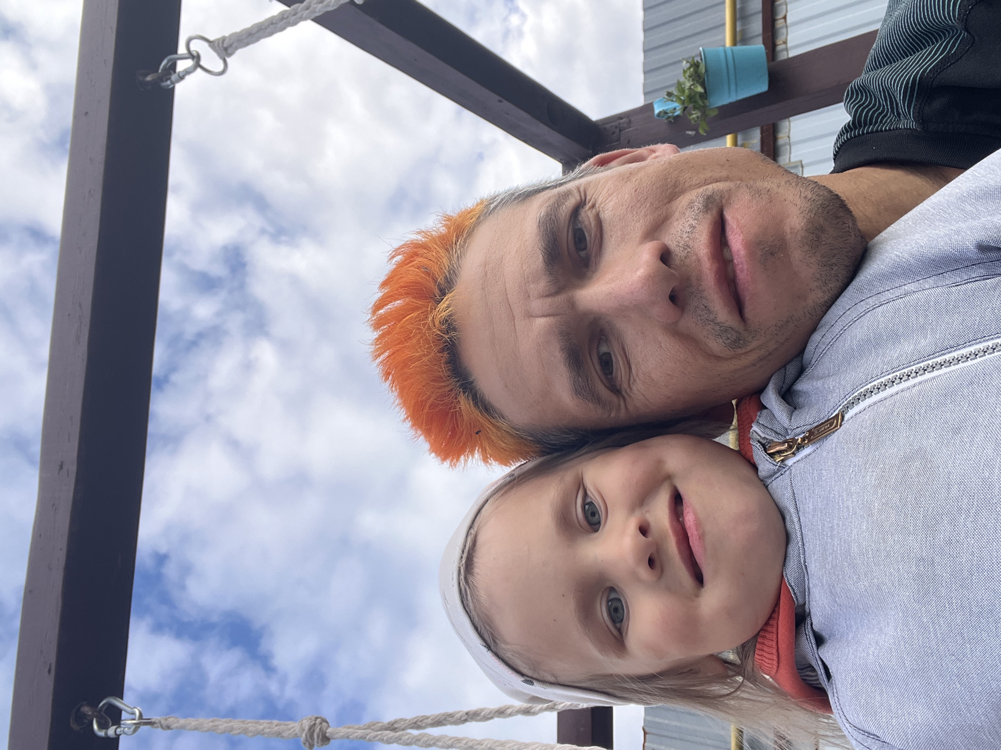 A selfie shows a man and a young girl on a swing set. The man has bright orange hair and is smiling. The girl is wearing a white headband and a gray jacket with an orange hood. They are both looking at the camera. In the background, there is a wooden swing set structure, a blue bucket with a plant, and a corrugated metal fence. The sky is cloudy. The location is identified as Chaikovsky, Russia.