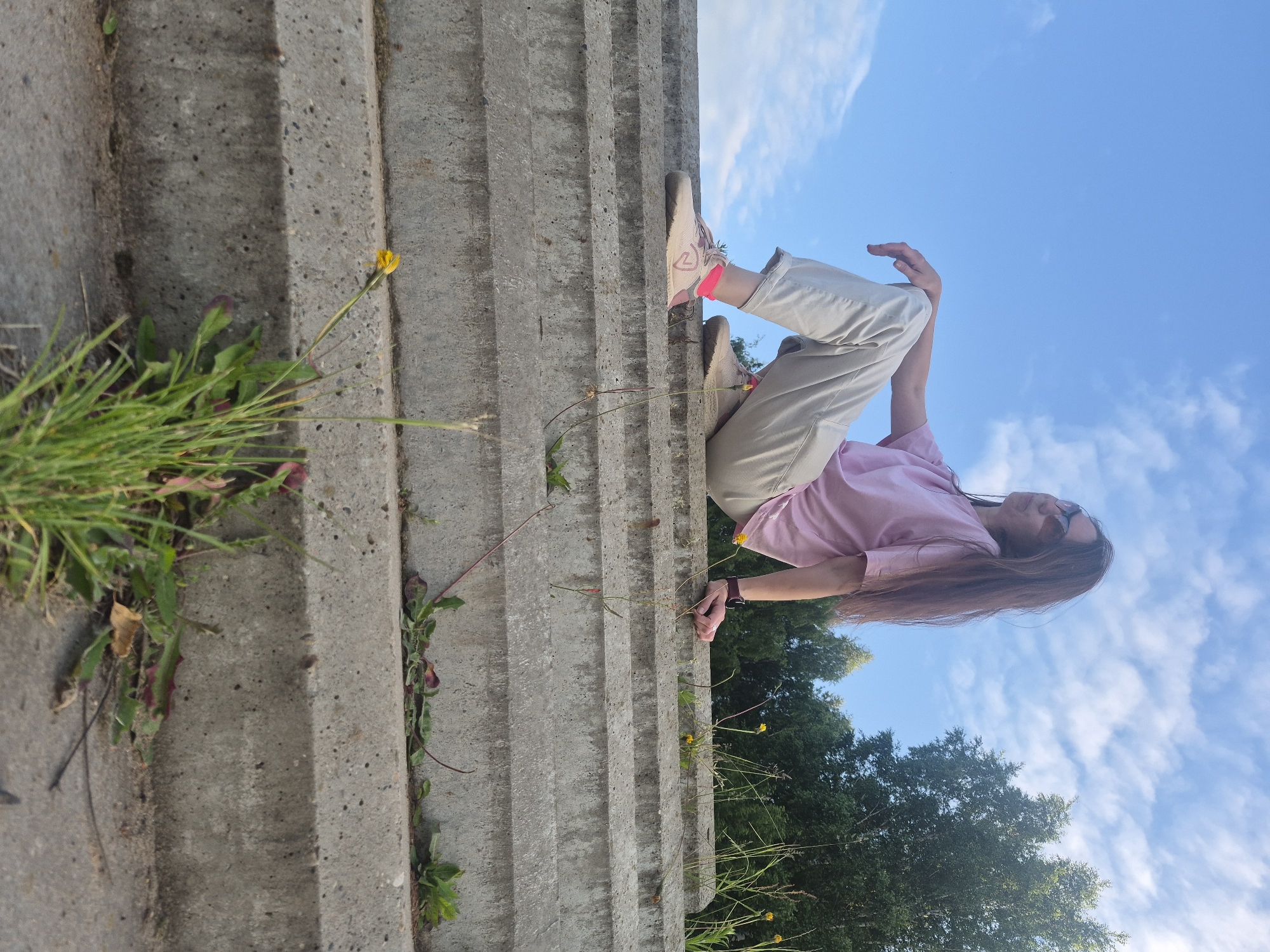 The image, taken in the city of Chaikovsky, Russia, presents a low-angle outdoor shot of a woman seated on concrete steps under a bright, cloudy sky.
A woman with long, dark brown hair and wearing sunglasses is positioned on the uppermost visible concrete step, facing slightly right and looking upwards with a subtle smile. She is dressed in a light pink short-sleeved t-shirt, light beige or off-white loose-fitting pants cuffed at the ankles, and light-colored athletic shoes accented with pink, complemented by bright pink socks. A dark-banded wristwatch is visible on her left wrist. Her left hand rests on her thigh, while her right hand is positioned on the concrete step behind her.
The foreground and mid-ground are dominated by several weathered concrete steps, exhibiting a coarse texture and minor surface irregularities. Various small plants, including clumps of green grass, weeds, and a few yellow dandelions, grow in the crevices and along the edges of the steps, particularly prominent in the lower left. In the background, beyond the woman and the steps, a clear blue sky with scattered white cumulus clouds is visible. A dense line of green deciduous trees occupies the upper right portion of the background, suggesting a wooded area. Additional sparse grasses and dandelions are present on the ground level behind the top step, adjacent to the trees. The scene is illuminated by natural light, indicating a sunny day.