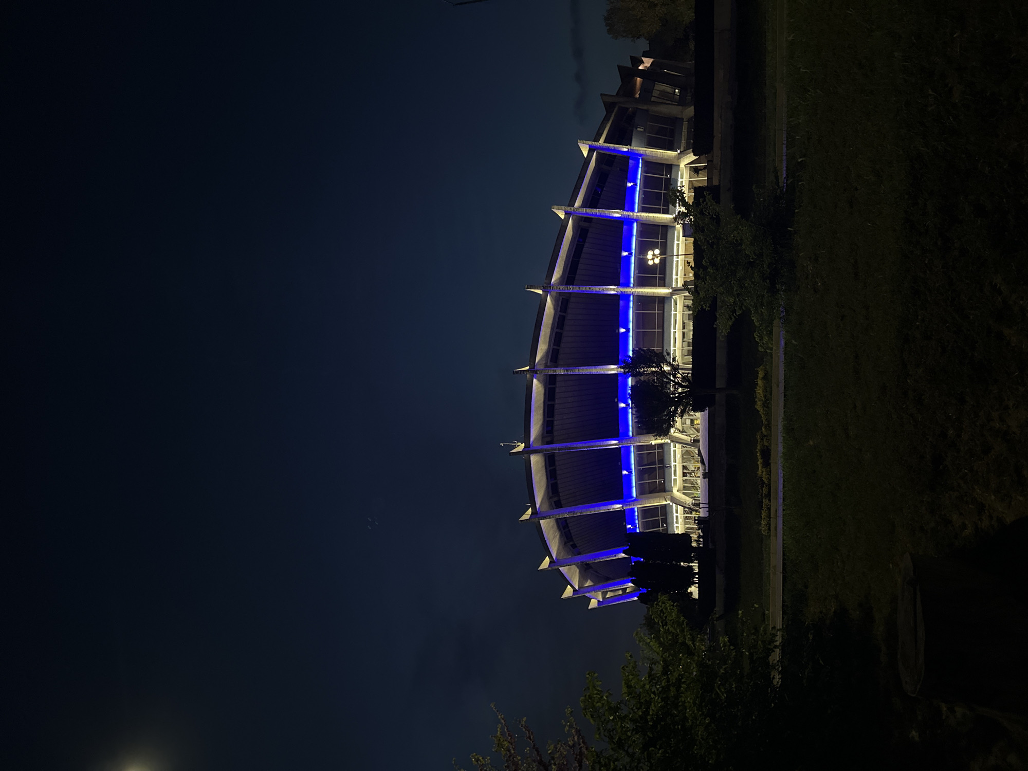 A night-time shot depicts the Palace of Culture and Sports in Varna, Bulgaria, featuring blue accent lighting. The building's exterior is visible against a dark sky. Lush greenery and landscaping flank the building on the right side of the image, while the left side shows an expansive dark area, likely the night sky above. The perspective gives a clear view of the building's distinctive architectural design, including its tiered levels and structural supports. No people are visible in the image.