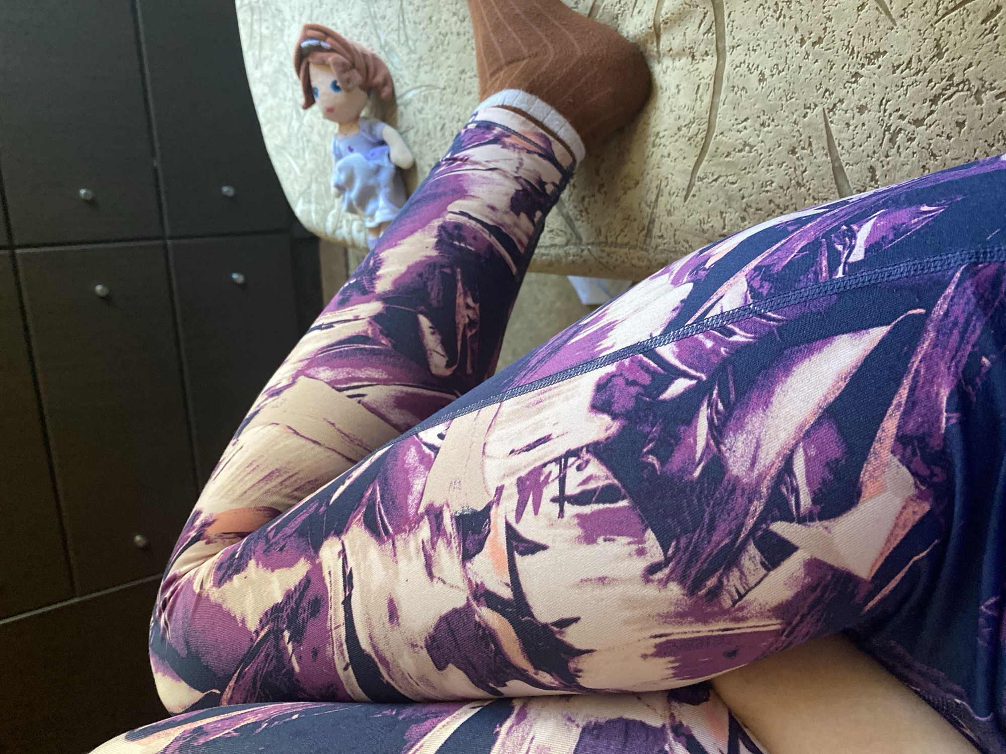 In an indoor setting, a person's legs are visible, wearing purple and cream patterned leggings and brown socks. The legs are bent, with the right leg resting on a textured surface. A small doll with brown hair and a blue outfit lies on the same surface. In the background, a dark brown dresser with silver knobs is partially visible. The environment appears to be a bedroom or living area, suggesting a domestic space. The lighting is soft, casting gentle shadows.