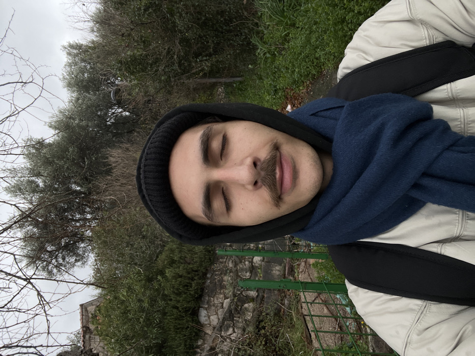 A male individual, appearing to be between 20 and 35 years old, is depicted in a frontal, waist-up outdoor portrait. He has medium skin tone, dark eyebrows, a prominent mustache, and his eyes are closed. He is wearing a black knitted beanie, a black hooded garment with the hood up, a dark blue scarf wrapped around his neck, a light-colored (likely beige or light grey) padded jacket, and a black backpack.
The background shows an outdoor environment consistent with a hillside or elevated natural area within the city of Budva, Montenegro. To the left, a stone retaining wall is partially covered by green leafy bushes. A section of a green metal fence or railing is visible at the lower-left. Above the wall, a part of a building with a reddish-brown tiled roof is discernible. To the right, dense green vegetation, including bushes and trees with both leaves and bare branches, fills the mid-ground and background. The sky is overcast and grey. A narrow path or ground area is visible at the lower right. The specific location within Budva is not identifiable by street or landmark, but the architecture and vegetation are characteristic of the region. The individual is stationary, with no specific actions or interactions depicted beyond posing for the photograph.