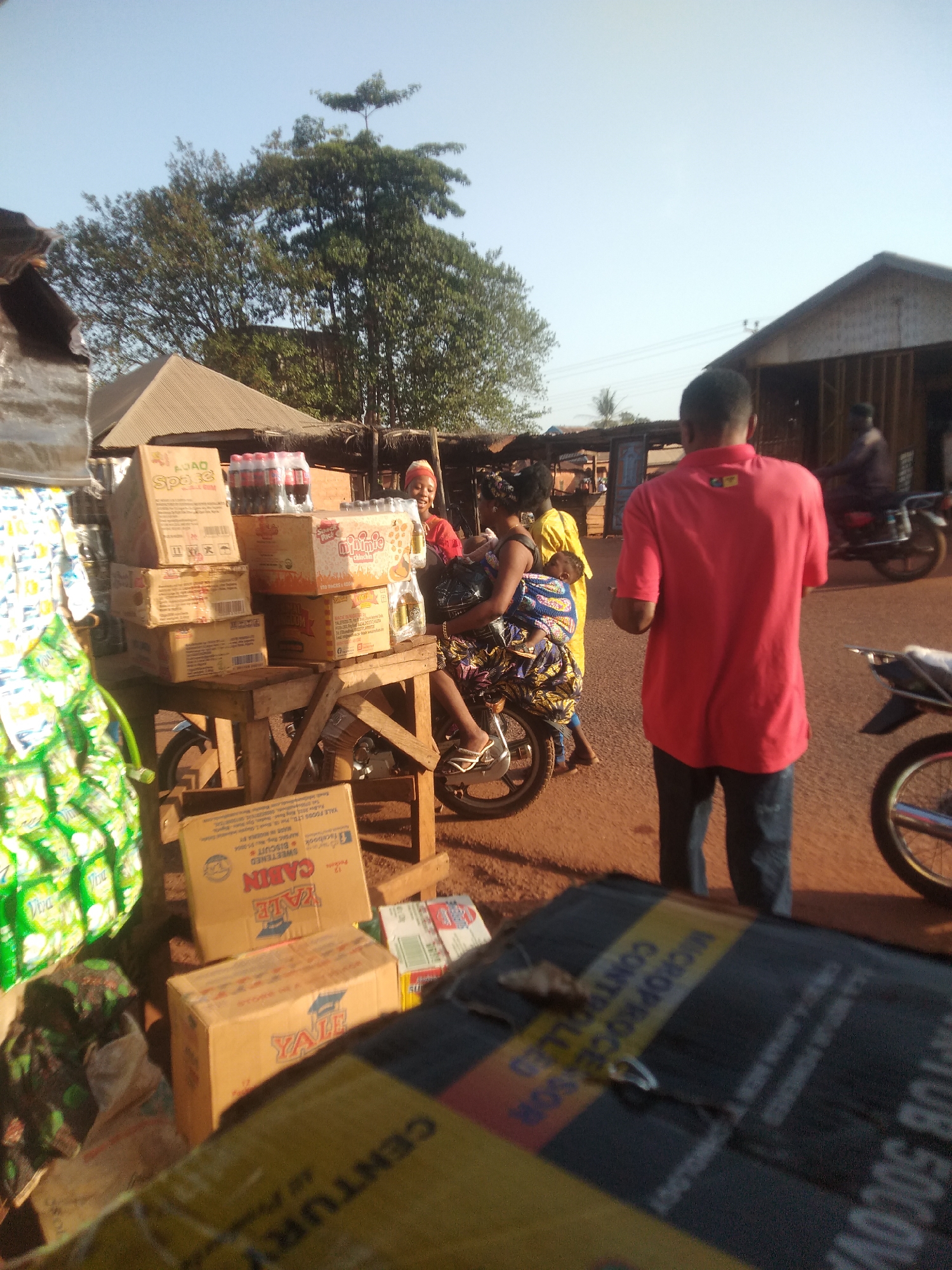 The image depicts a street scene in Ejule, Nigeria. At the center, a woman wearing a patterned dress and sandals is seated on a motorcycle, carrying a child on her back. A woman with red head gear is standing next to the motorcycle. Boxes of various goods (e.g., 'Space Chinchin', 'Yale Cabin biscuit', 'Minimie chin-chin') are stacked on a wooden table supported by a motorcycle. The background includes a man in a red shirt with his back to the camera, other people, buildings, and a tree. Boxes of products are displayed at the foreground of the image.