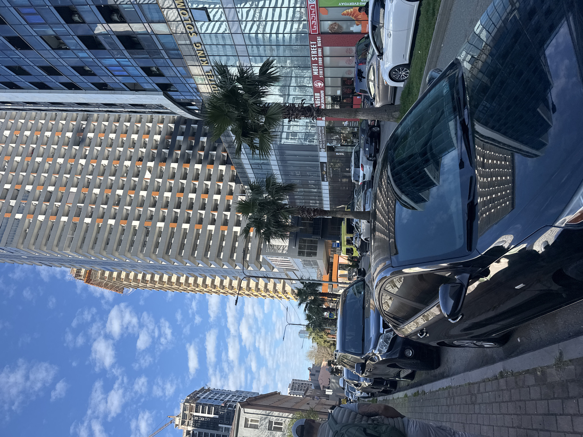 This image captures a vibrant urban street scene, likely during the daytime, under a bright blue sky dotted with scattered white clouds. The setting appears to be a commercial district characterized by modern architecture and palm trees, suggesting a warm climate.
The scene features a row of parked cars along the right side of a sidewalk, including dark-colored sedans and a white car. In the foreground on the bottom right, a person is partially visible, wearing a hat and what appears to be a jacket, possibly walking or standing.
Dominating the background are tall buildings. On the left, a multi-story building displays a distinctive facade with repetitive rectangular balcony or window structures. To its right, a sleek glass-clad building reflects the sky and surrounding elements. This glass building prominently features the text "HOTEL KING SOLOMON" vertically along its side. Other visible text includes "MOFI STREET" with "STREET FOOD - DONE RIGHT" below it, "KING & QUEEN SALON," and "ING EVERYDAY." Some signage also appears in Georgian script, including "საჭმელი" (Sachmeli, meaning food) and "JUICES." Numbers like "3063 & 5006," "1560," and "00904" are also present on various storefronts.
Several palm trees line the street, interspersed between the parked vehicles and the buildings. In the distance, on the lower left, another building appears to be under construction, indicated by visible scaffolding and unfinished elements. The overall impression is one of a busy, developing city environment.
