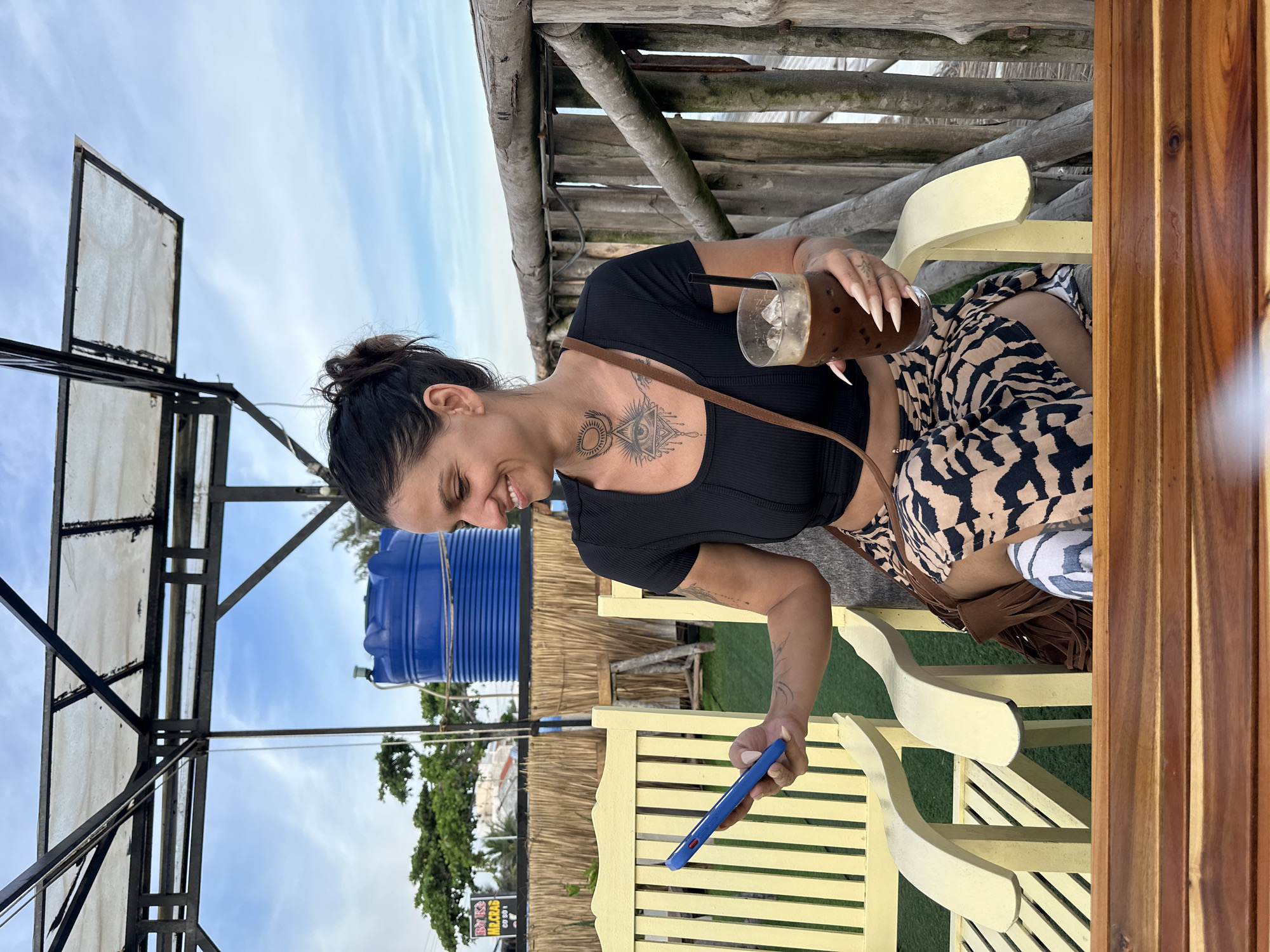 A woman sits at a wooden table outdoors, holding a drink and a blue smartphone. She is wearing a black top and a patterned skirt, with a tattoo visible on her chest. The setting appears to be a casual eatery or cafe, with a wooden structure overhead and yellow chairs. In the background, a large blue water tank and lush greenery are visible under a partly cloudy sky. The overall impression is one of a relaxed, daytime scene, possibly in a tropical or coastal location, given the vegetation and open-air setting. The woman seems content, with a slight smile on her face as she looks at her phone. Visible text includes "MR. CRAB" on a sign in the background.
