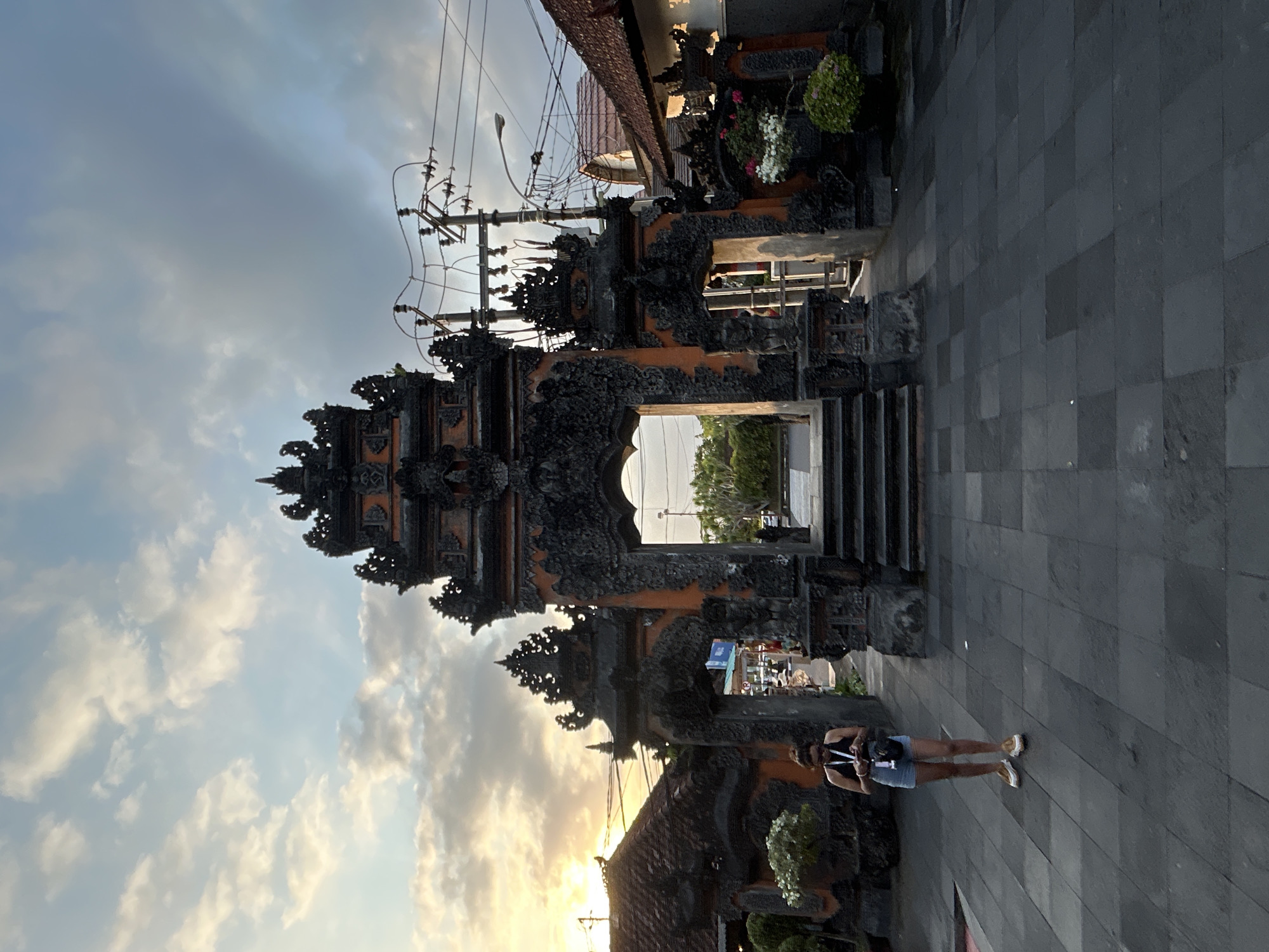 A Balinese temple gateway, likely in Kediri, Indonesia, is captured in an outdoor, low-angle shot during twilight. The ornate gateway features intricate dark stone carvings against an orange backdrop, with steps leading up to a central archway. The sky above is a mix of blue and cloudy hues, with the setting sun casting a warm glow on the left side of the frame. A person, appearing to be a woman, stands in the foreground on the left, wearing a dark tank top, denim shorts, and yellow sneakers, holding a phone. The ground is paved with dark square tiles. On either side of the gateway, traditional tiled roofs of nearby buildings are visible, along with potted plants. Electrical wires and a lamppost are also present in the background. The overall scene suggests a cultural landmark in a tropical urban environment.