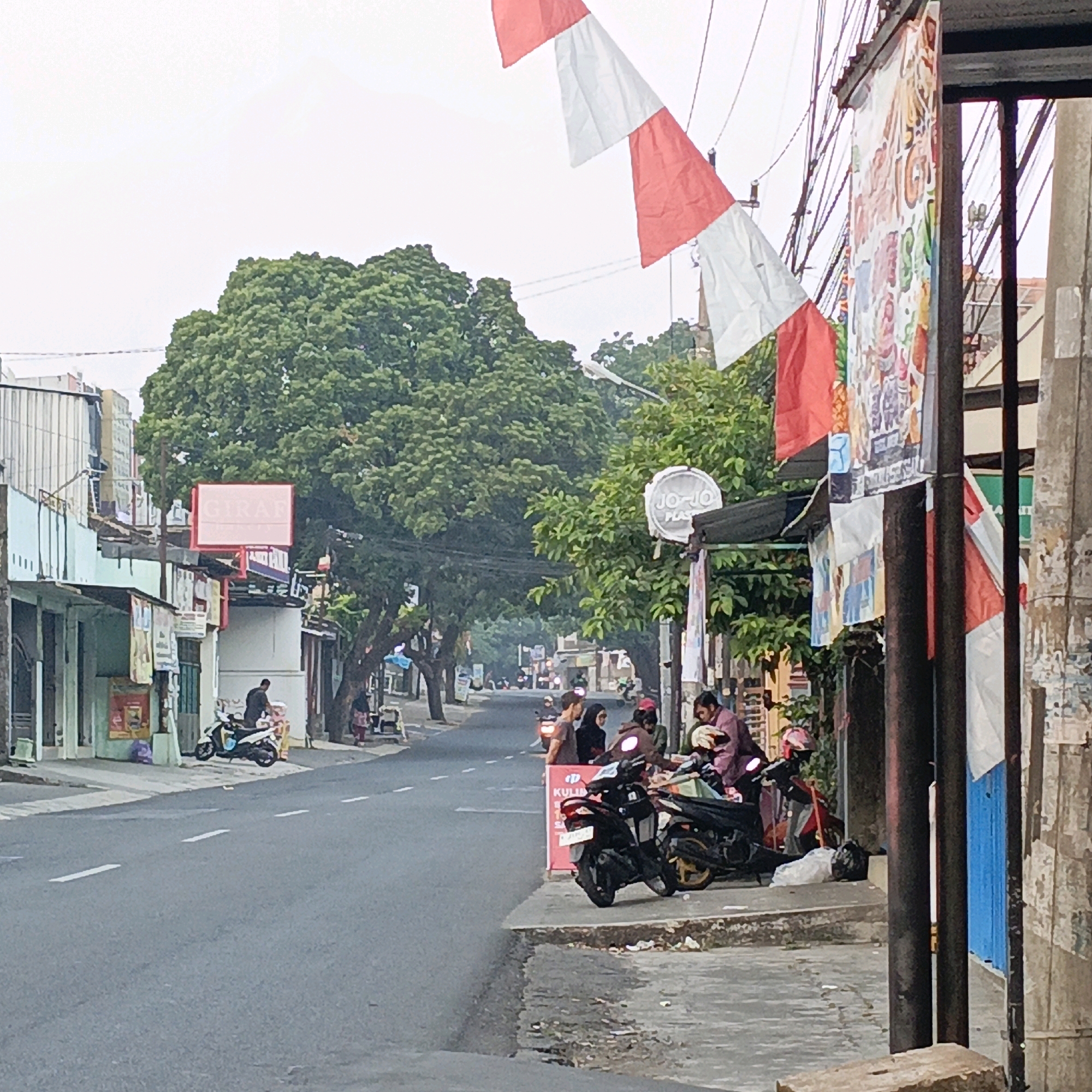 The image shows a street scene in Salatiga, Indonesia, with a focus on a row of shops and people gathered along the sidewalk. Red and white Indonesian flags are strung across power lines, suggesting a national holiday or celebration. Several motorcycles are parked on the sidewalk, indicating a common mode of transportation in the area. People are seen standing and sitting, some appearing to be patrons of the shops or vendors. The shops themselves have a variety of signage, including one that reads "GIRAF" and another with a "JO-JO PLAS" logo. The street is paved and appears to be a typical urban road with lane markings. The overall impression is of a bustling street with everyday life unfolding.