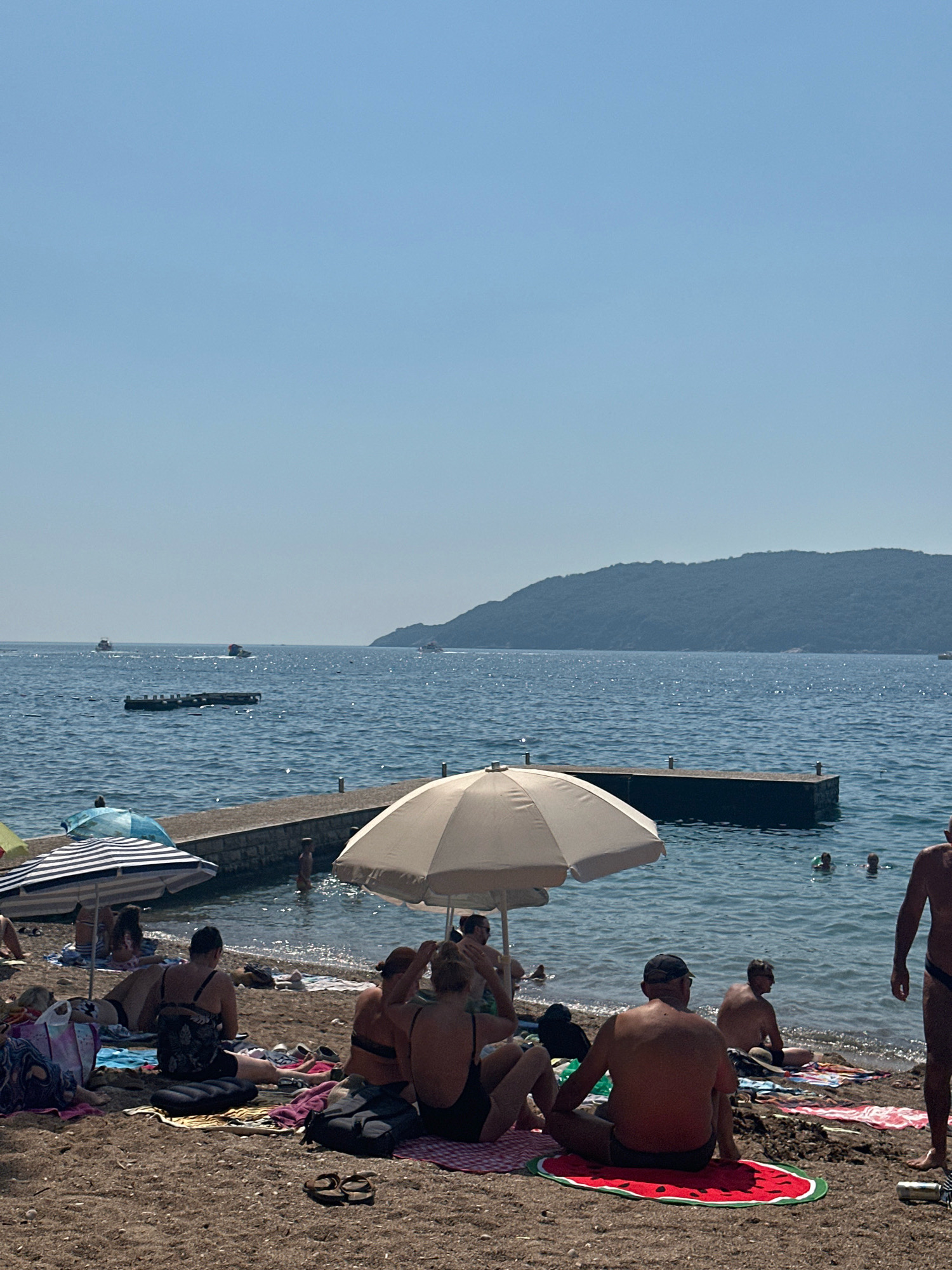 This is a vertical, eye-level, outdoor, wide-angle shot of a crowded beach in Budva, Montenegro, on a clear, sunny day. The foreground features a sandy beach with several people sunbathing and sitting on towels and mats. In the mid-ground, there is a body of water with a small concrete pier extending into it. Several small boats are visible on the water, and in the background, a large, tree-covered hill forms the coastline.
In the foreground, a variety of beachgoers are depicted. On the left, a person in a black swimsuit sits with their back to the camera, facing the water. To their right, a group of people are seated on towels. One person, in the center of the frame, wears a black swimsuit and rests their chin on their knees. To their right, two individuals, one in a black bikini and the other with their back to the camera, are also seated. Further right, a man in a black baseball cap and swim trunks sits cross-legged on a red, watermelon-patterned mat. Another man, partially visible on the far right, stands wearing black swim trunks. Beach umbrellas, towels, and bags are scattered across the sand.
The water appears calm, with gentle waves lapping at the shore. A few people are swimming or wading in the water near the pier. The sky is a clear, light blue, with no clouds visible. The overall atmosphere of the image is one of a busy summer day at the beach.