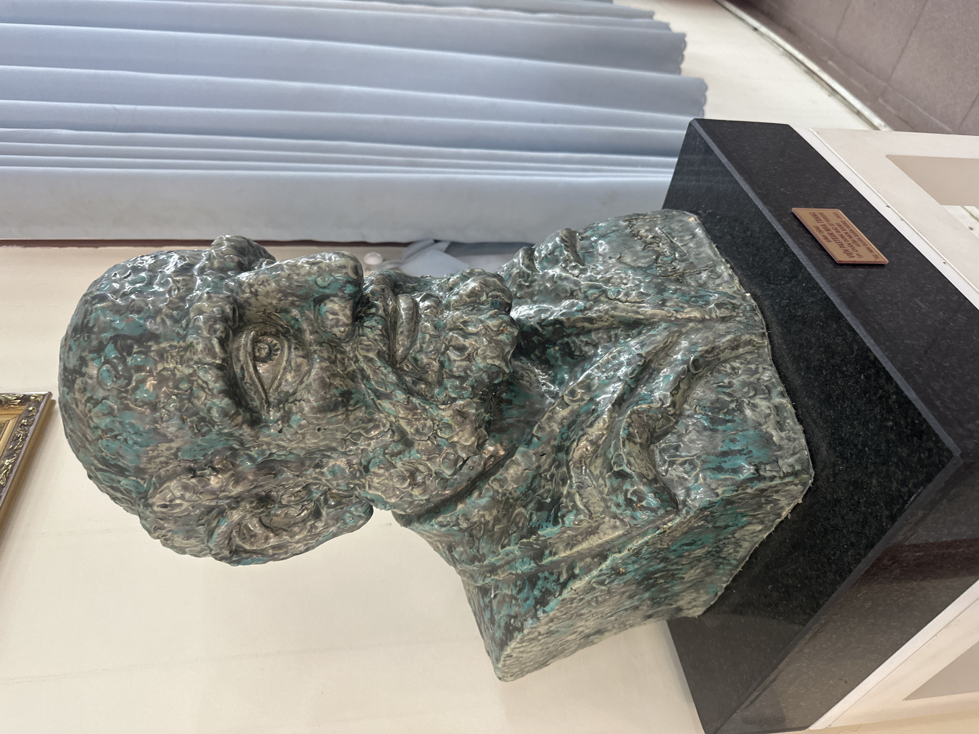 The image shows a bust of a man with a weathered, textured metallic surface, exhibiting shades of green and gray. The bust is placed on a black marble pedestal. A plaque on the front of the pedestal reads: "VIỆN PASTEUR NHA TRANG TP. QUẬN DỊCH BẮC SĨ VERSAILLES GÔM MÔN XUÂN HƯƠNG - 2023". This translates to: "INSTITUTE PASTEUR NHA TRANG CITY. EPIDEMIOLOGY DOCTOR VERSAILLES GÔM MÔN XUÂN HƯƠNG - 2023". The background features a wall and a light blue curtain. The lighting appears to be indoor ambient light, casting soft shadows.