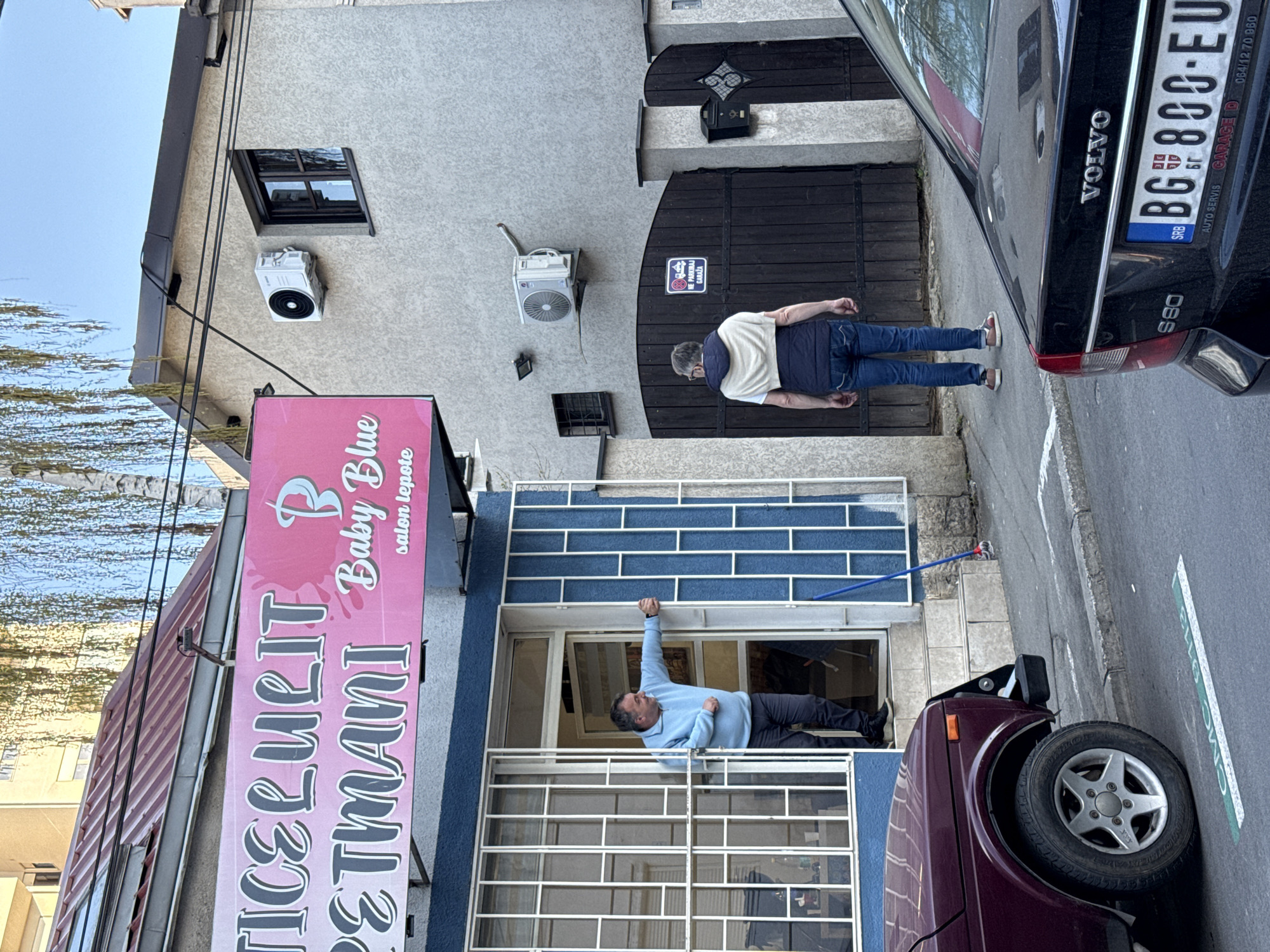 The image shows a street scene, seemingly in Belgrade, Serbia, with a building housing a beauty salon called "Baby Blue salon lepote". A sign next to it reads, "ICELULIT PETMANI". A man is seen inside the salon window, appearing to be cleaning or fixing something. Another man is standing on the sidewalk near a black Volvo S80 with the license plate "BG 800-EU", indicating Serbian registration. A sign on the building reads, "NE PARKIRAJ GARAŽA", which means "Do not park Garage" in Serbian. A dark purple/burgundy car is parked near the salon window, partially blocking the sidewalk. There are also air conditioning units on the building's exterior.