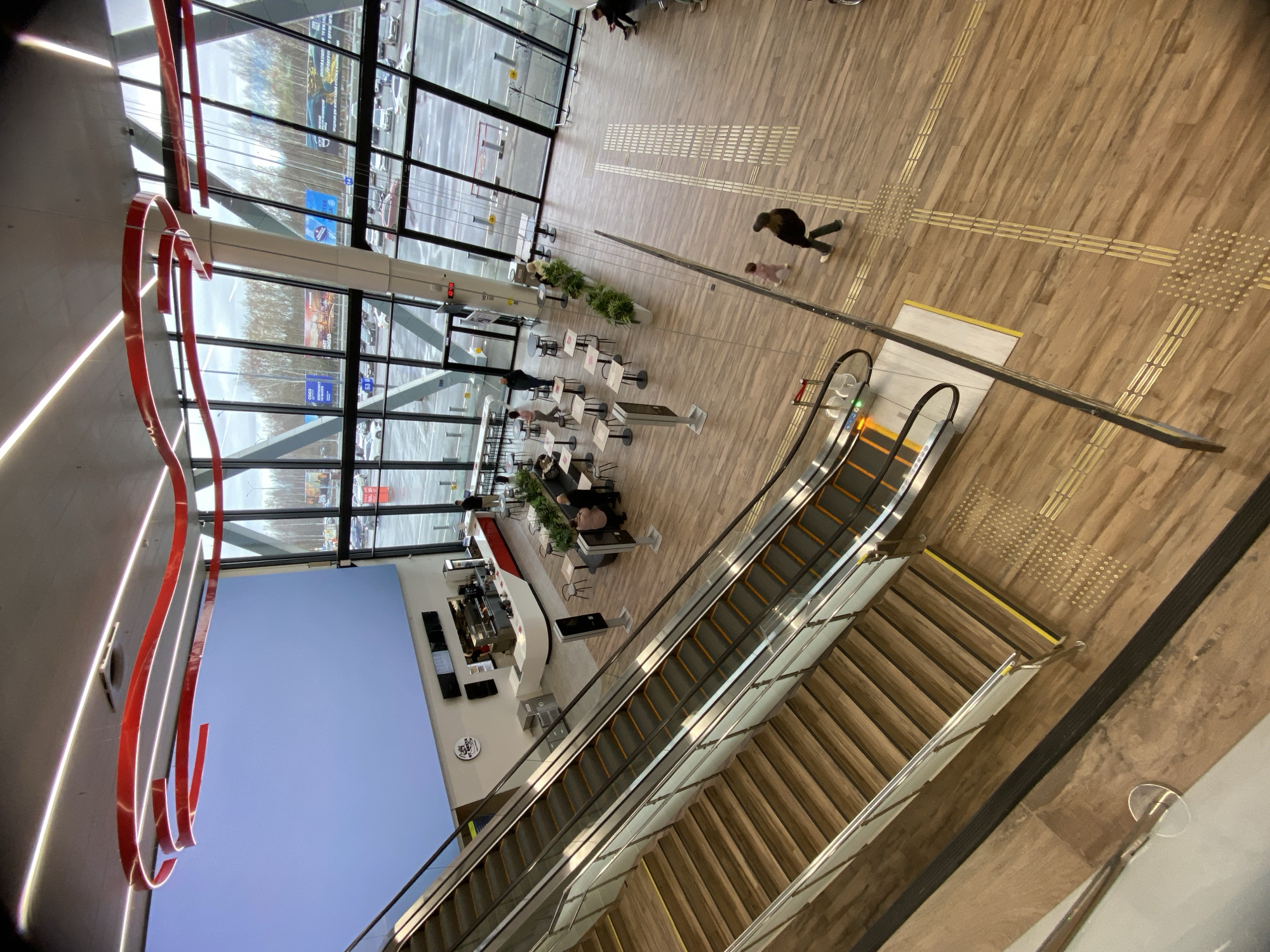 An overhead, slightly angled view captures the interior of a spacious, modern public building, likely a shopping center or transport hub. The floor is covered in light-colored wooden planks, featuring tactile paving strips and dots for orientation. Large, full-height windows on the left reveal an overcast outdoor scene with parked cars.
In the center-right, a long escalator descends, paralleled by a wooden staircase. Several individuals populate the scene. One person in a dark jacket walks across the wooden floor with a small child in a pink outfit. Further left, a cafe area is visible with multiple tables and chairs, where other people are seated. Green potted plants decorate this space.
A distinctive red, wavy architectural or artistic element runs along the upper left wall. A large, solid light blue panel covers a section of the lower left wall.
Visible text includes "COCO" on a blue sign and "ОКНА РОСТА" (OKNA ROSTA) with smaller text "ПОЛНЫЙ КОМПЛЕКС УСЛУГ" (FULL RANGE OF SERVICES) seen through the windows. A circular white logo with a dark crest and faint text is on the cafe wall, and "ЦЕНТРАЛЬНЫЙ" (Central) is partially legible on another sign nearby. The lighting suggests daytime, with diffused light entering from outside.