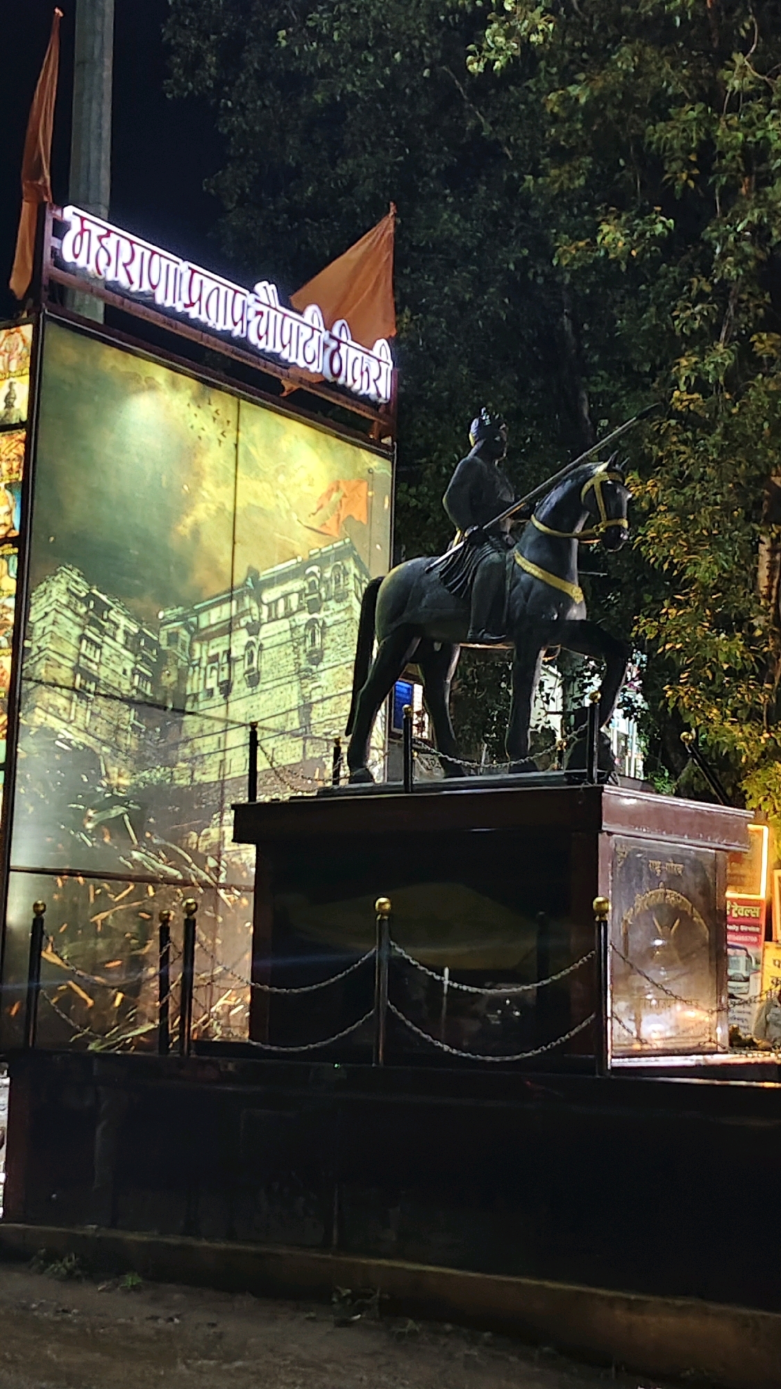The image shows a statue of a man on horseback at night in Thikri, India. The statue is dark in color and depicts a figure in historical attire holding a spear. The horse is also dark and appears to be in motion. Behind the statue is a large illuminated mural depicting a historical building with an orange flag. A neon sign with white lettering in an Indian script is mounted above the mural. Two orange flags are visible on poles to the left and right of the sign. The statue and mural are enclosed by a low barrier with posts and chains. Trees with dark foliage are visible in the background, and the ground in front of the statue appears to be a dirt or gravel surface. The overall scene is dimly lit, with the light primarily coming from the illuminated mural and sign.