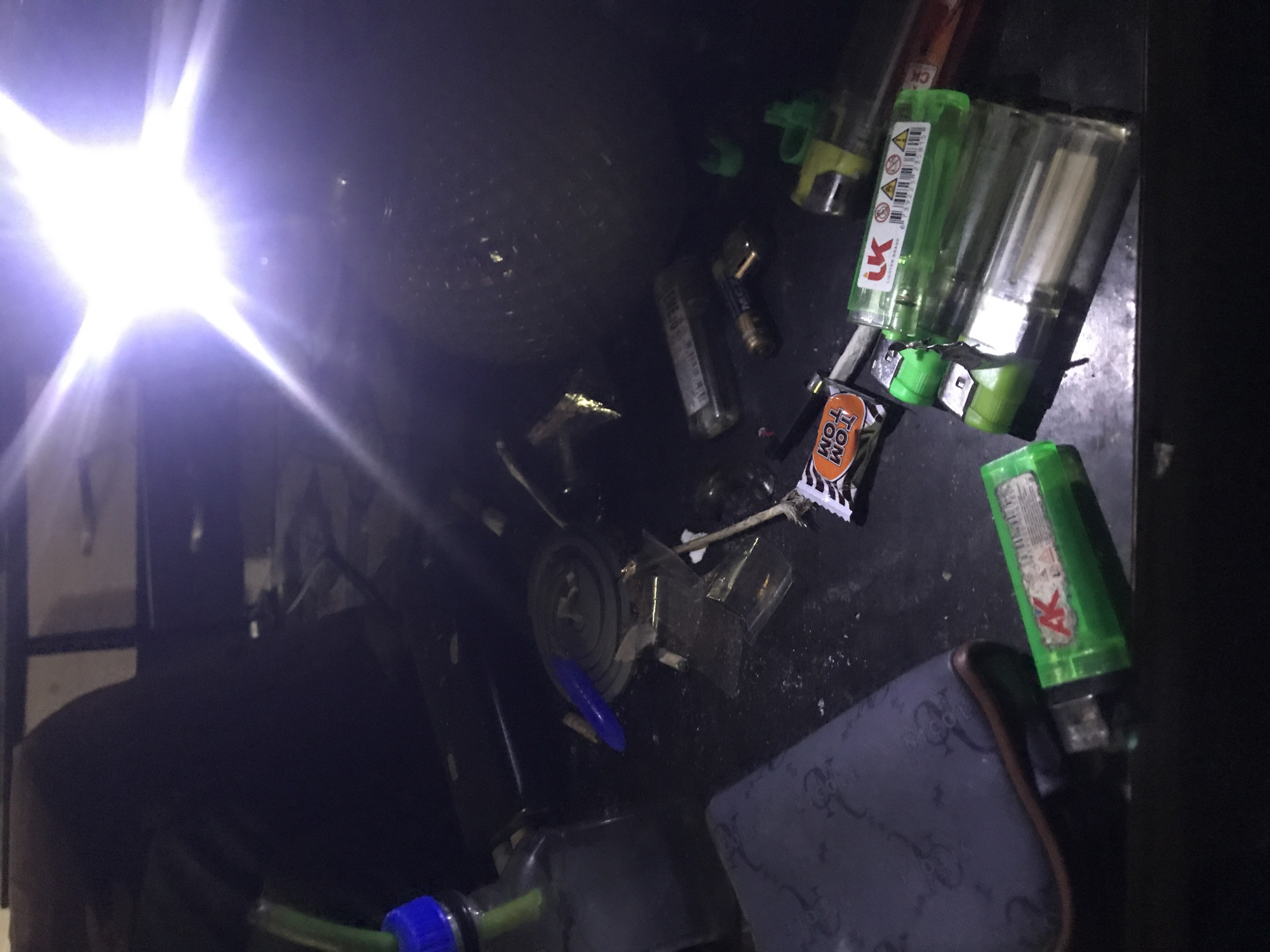In an indoor, dark environment, a collection of drug paraphernalia is scattered across a flat surface. A glass bottle with a green tube and blue cap is visible on the left, possibly a bong. Nearby, several lighters of various colors, including green, clear, and red, are present. Small pieces of what appear to be glass or plastic are also spread out. Additionally, a dark gray object with a patterned design, resembling a pouch or case, rests on the left side. A few rolled items, potentially cigarettes or joints, are also discernible amongst the debris. The scene is illuminated by a bright light source, likely a flash, which casts strong shadows and highlights.