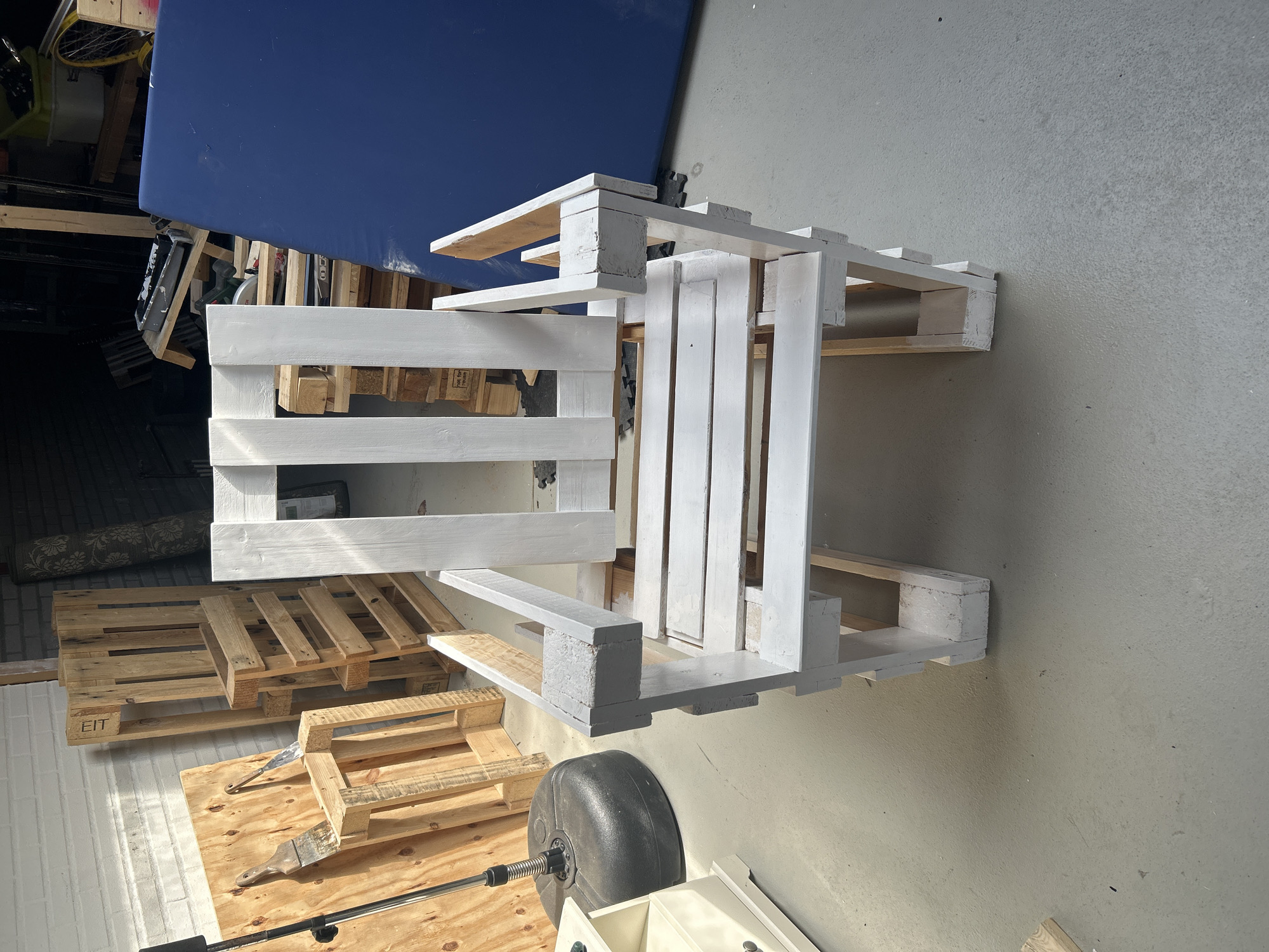 A white, unfinished chair made from pallet wood sits in the center of the frame. The chair has a high back with three vertical slats and armrests. The seat is made of horizontal slats. The chair is constructed from rough, unvarnished wooden pallets, some of which are still visible in their original state. The chair's construction suggests it is a DIY project. To the left of the chair, a black, weighted base of what appears to be a floor lamp or a stand is visible, along with a light-colored cabinet. Behind the chair, several more wooden pallets are stacked, some upright and some on their sides. A large, bright blue tarp or mattress covers a significant portion of the background to the right. The floor is a light gray concrete. The scene appears to be in a workshop, garage, or outdoor covered area, possibly a carport, given the presence of unfinished materials and tools that might be implied by the context of pallet furniture construction. The location is in Lisse, Netherlands.