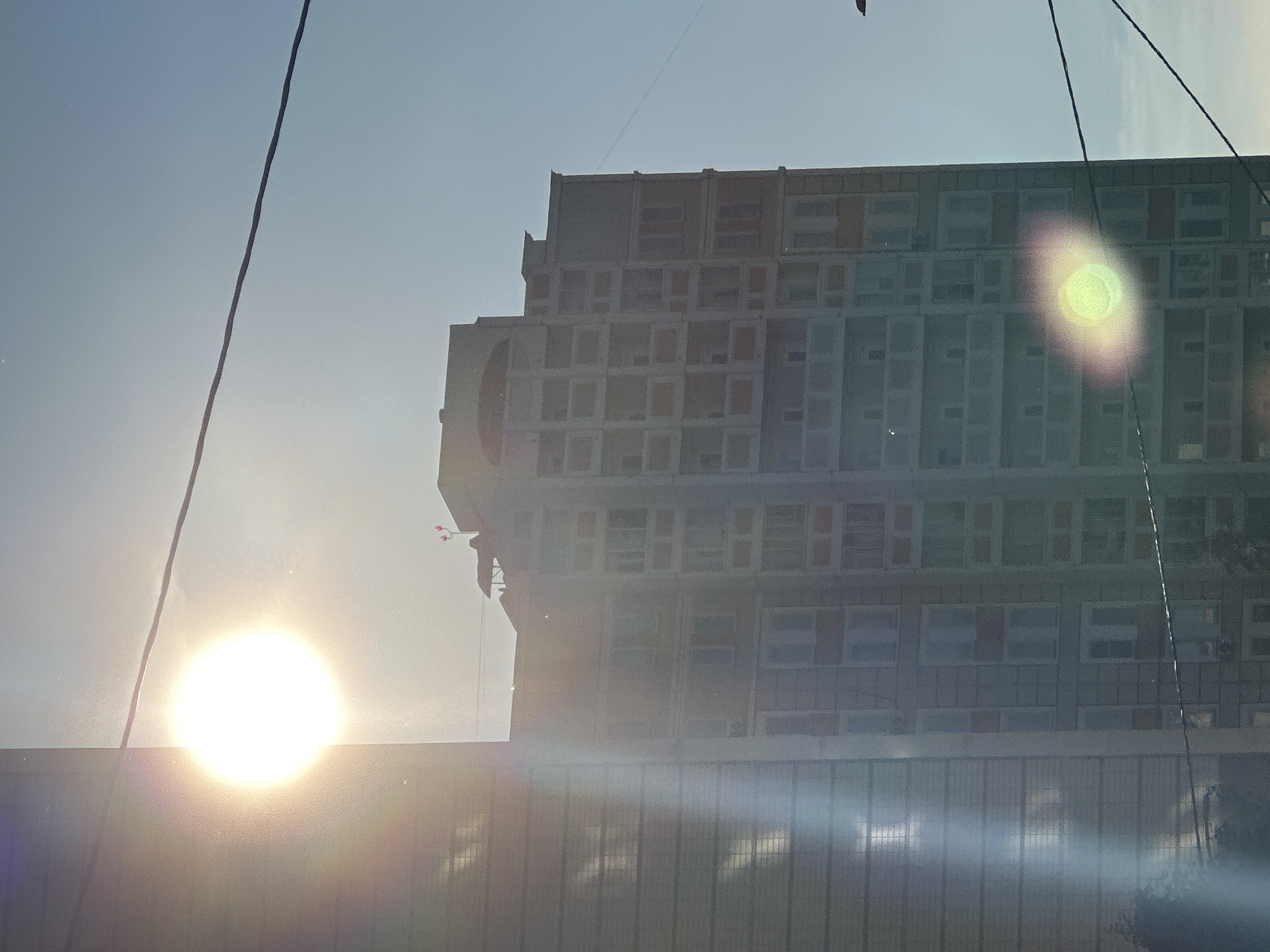 The image depicts a tall, multi-story apartment building in the city of Podolsk, Russia. The sun is intensely bright and positioned in the upper left quadrant, causing lens flare that creates a large, circular, bright white and rainbow-colored halo effect. A portion of a neighboring building, also tall and with a reflective facade, is visible on the far left. The sky is clear and blue. Several black electrical wires are stretched horizontally across the frame, one in the upper portion and another lower down. The apartment building itself appears to be constructed from concrete panels with numerous windows and balconies. At the very top of the building, a small red and white weather vane or similar structure is visible. The overall scene is urban, under bright daylight conditions.