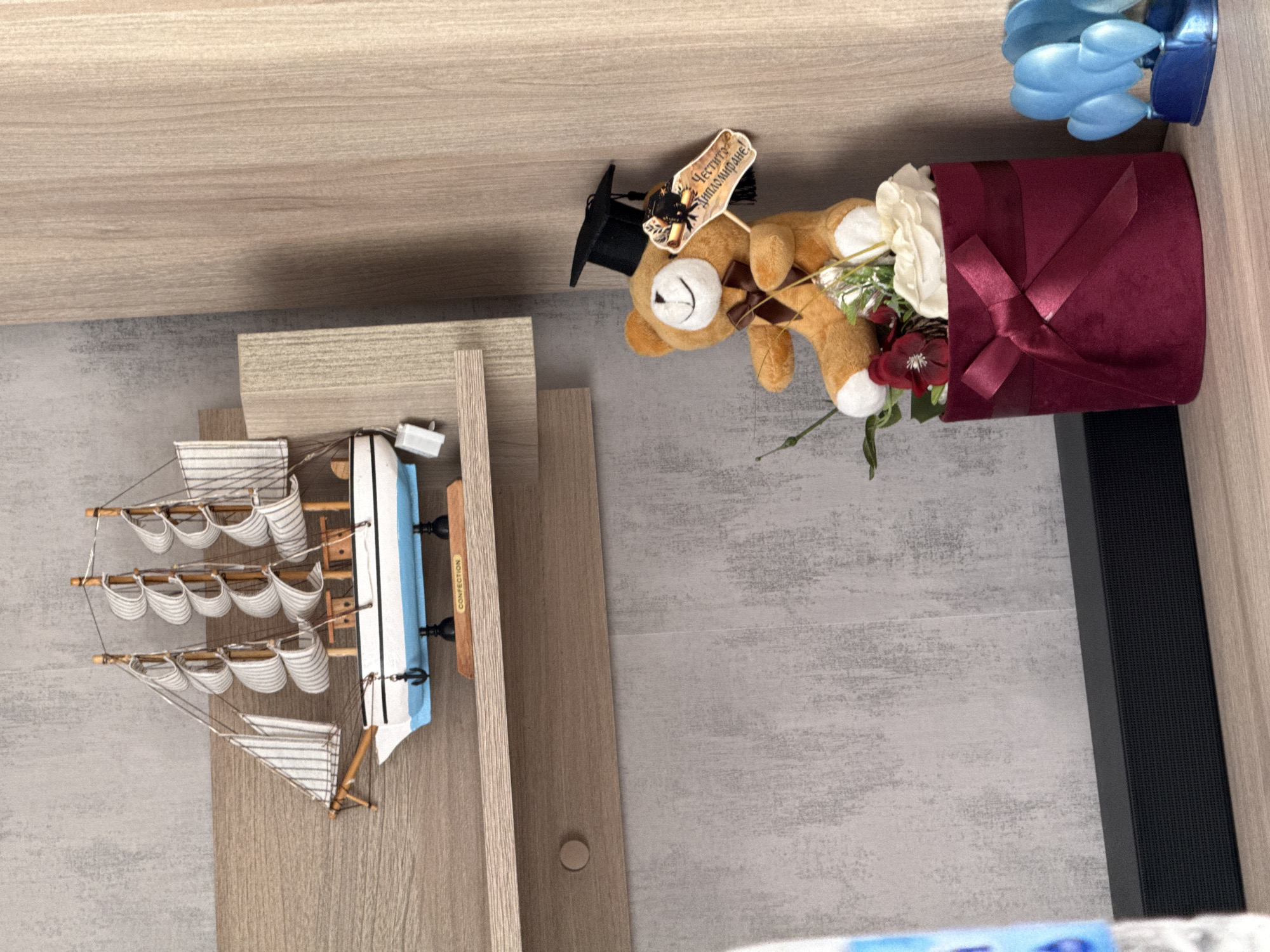 A high-angle, medium shot depicts a collection of items resting on what appears to be a light-colored wooden or laminate surface. The background consists of a gray, textured surface, possibly a wall or floor, with a dark vertical object to the right.
Arranged from left to right, the items include a model sailboat with white sails and a blue hull, positioned on a small, tiered wooden platform. Next to the boat lies a gift arrangement consisting of a graduation-themed teddy bear wearing a black cap and holding a scroll that reads "Честито Дипломиране" in Bulgarian, nestled inside a deep red box with a matching ribbon, accompanied by white roses and red flowers. To the far right are a few blue decorative objects, partially cropped out of the frame.
The image does not provide specific geographical data, therefore, the city of Lovech, Bulgaria, cannot be verified as the location.