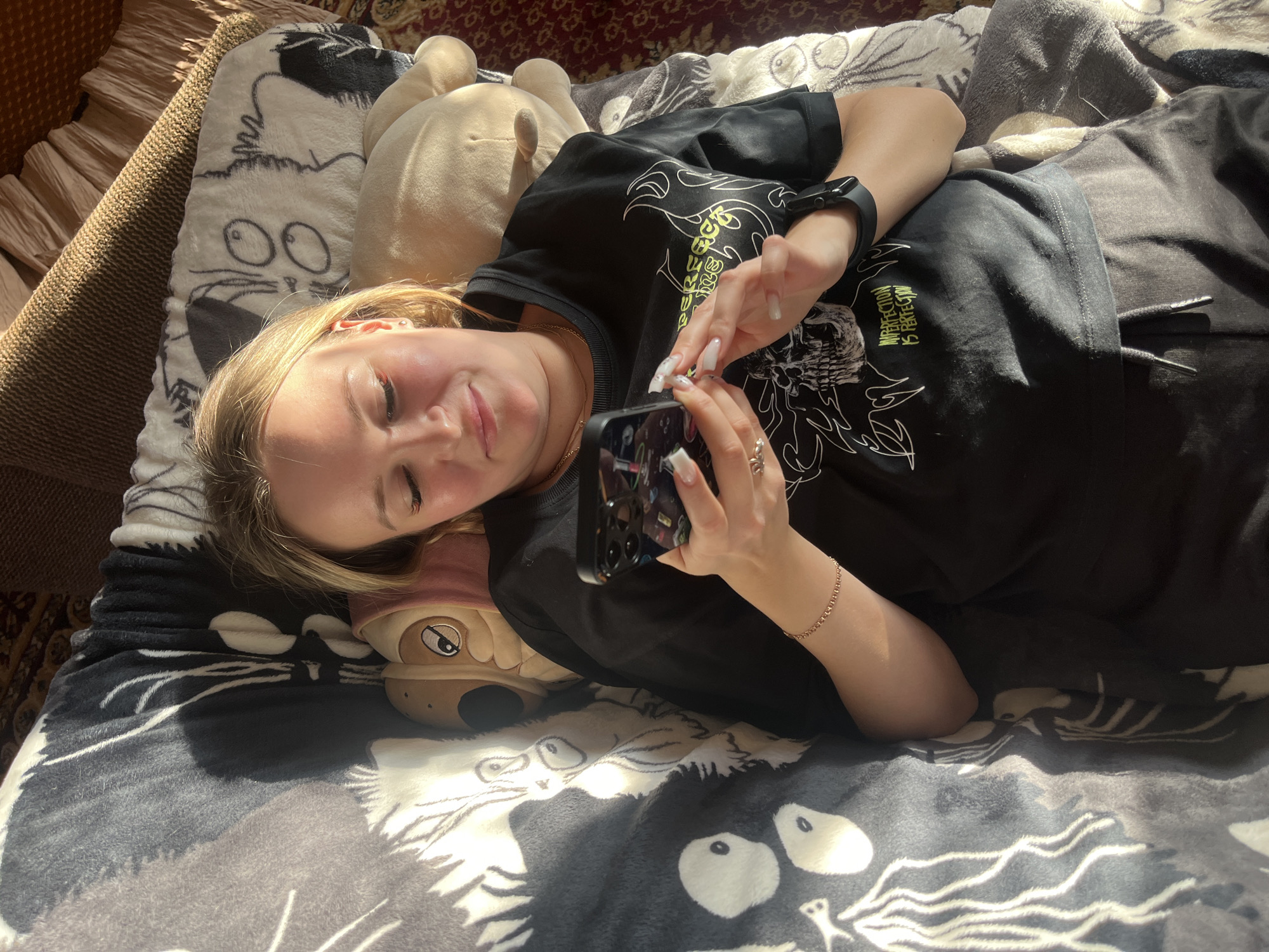 A young woman is lying on a bed. She is wearing a black t-shirt and black pants. She is holding a black smartphone in her hands. She is looking at the phone and smiling. A plush dog toy is next to her. The bed has a gray and white blanket with cartoon-like cat faces. The location is within the city of Podolsk, Russia.