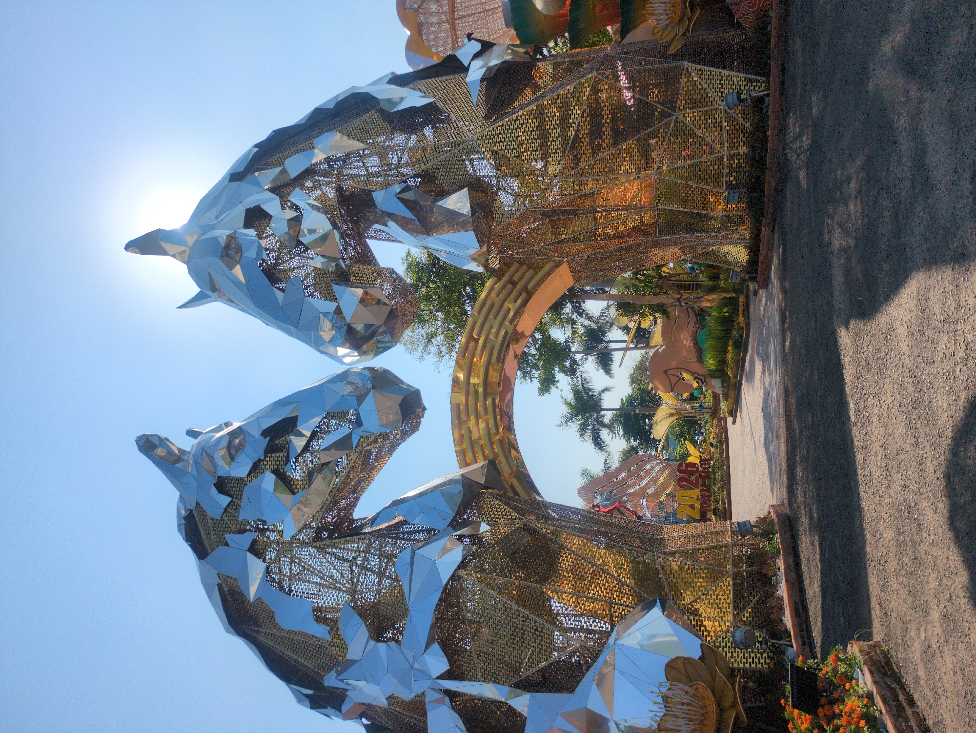The image shows an outdoor art installation in Tuy Hòa, Vietnam. Two large, mirrored horse heads are prominent, constructed from geometric shapes and wire mesh. A circular archway is positioned between the horse heads, leading to a path with trees and other art pieces in the background. The scene is brightly lit, likely during the day, with a clear blue sky. The walkway appears to be paved with asphalt, bordered by vegetation and flowers.
