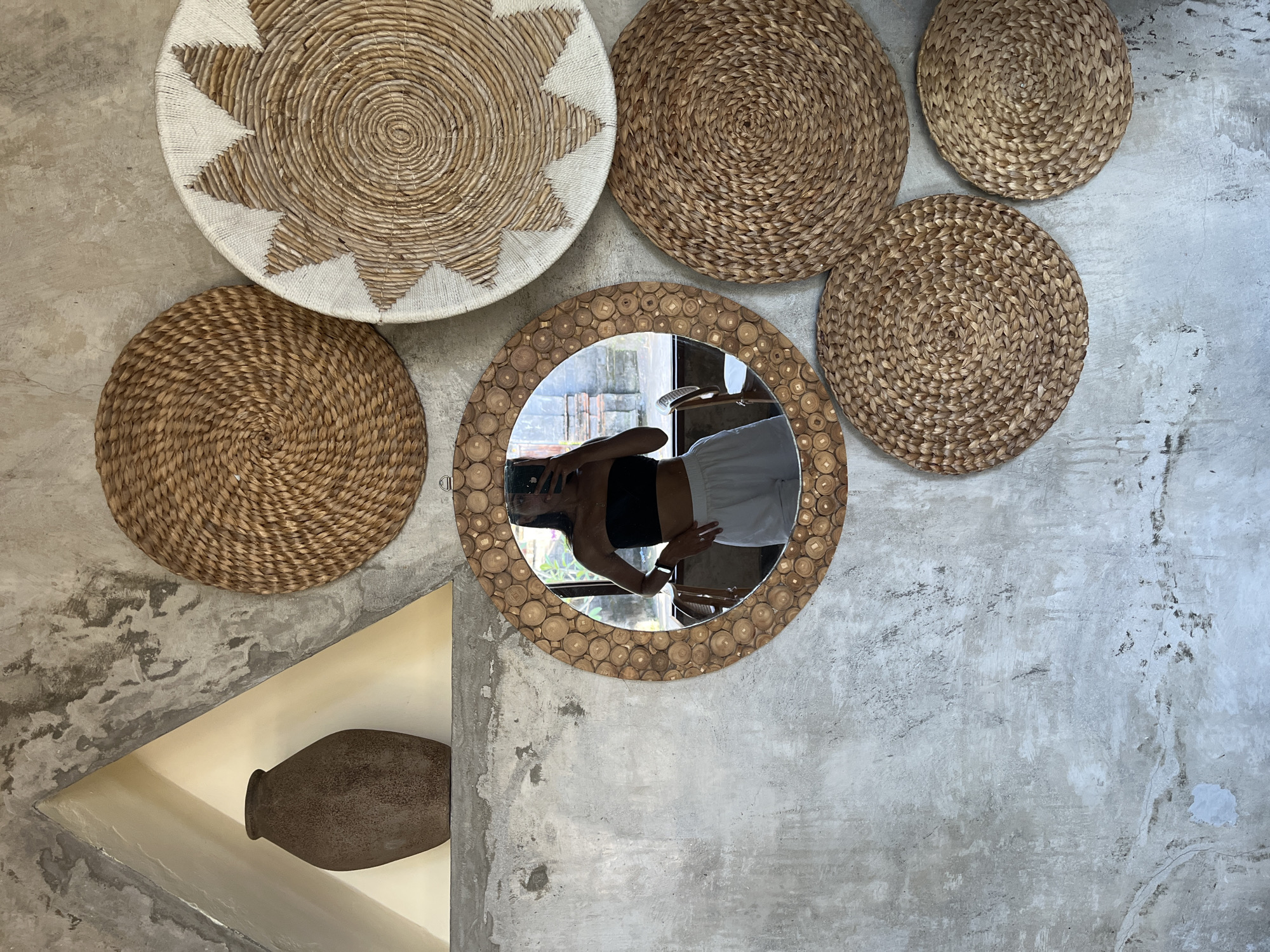 A scene inside a room in Sukawati, Indonesia, features a concrete wall decorated with woven circular mats. The arrangement includes a sun-shaped mat with white and tan patterns, several circular mats of woven reeds in varying shades of brown, and a round mirror with a wooden frame composed of cross-sectional wood slices. The mirror reflects a person taking a selfie, wearing a black top and light-colored shorts. To the lower left, there is a triangular niche in the wall housing a dark, bulbous vase. The overall impression is one of rustic decor.