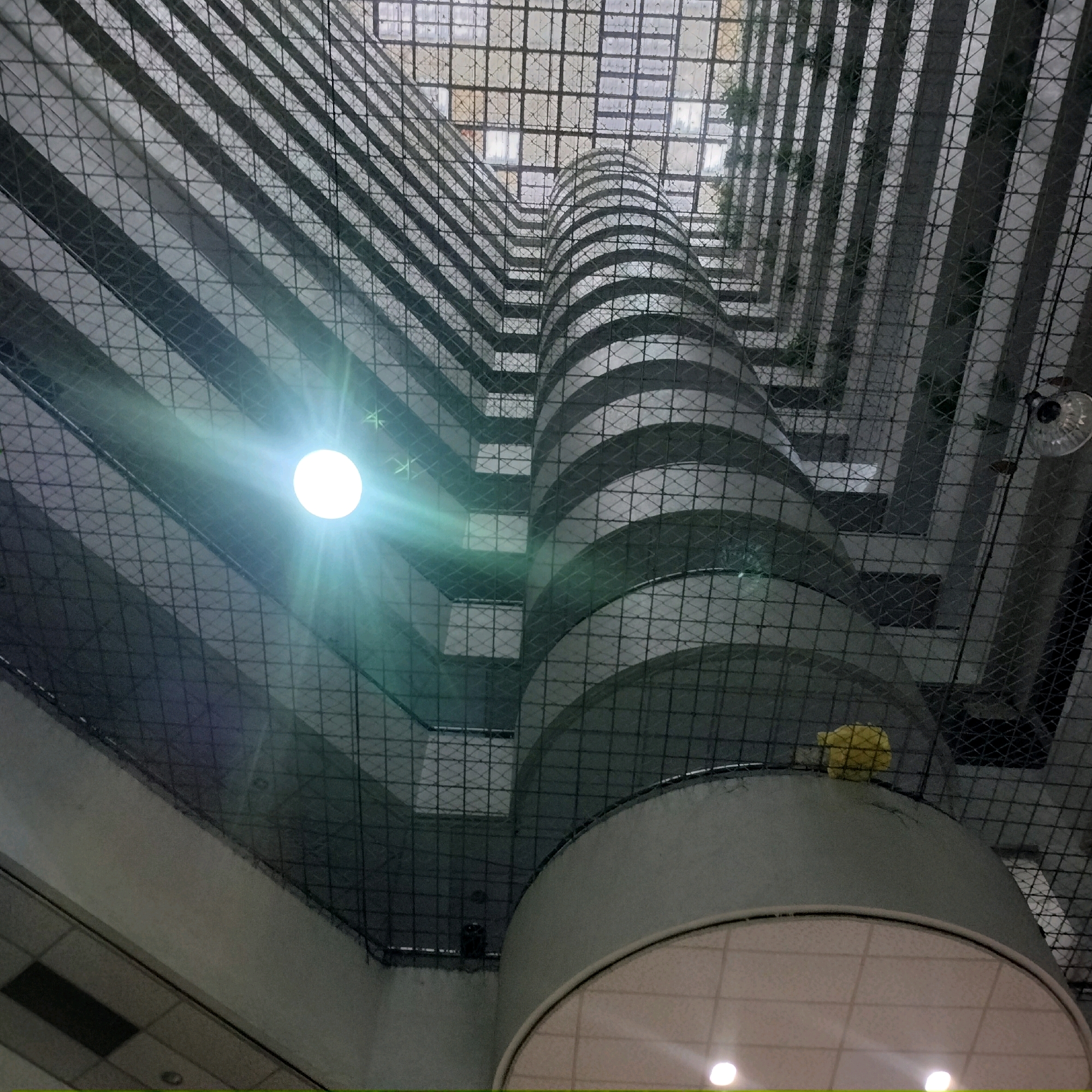 The image shows the interior of a large, multi-story atrium building with a glass roof and a central, cylindrical structure. The atrium is enclosed by tiered levels, each with a mesh safety barrier. The architecture features a spiral design. A bright light source is visible on the left side of the image, causing a lens flare and illuminating the scene. On the right side, a light fixture is attached to a support structure. A small, yellow object, possibly a bag, is placed on the edge of the central cylinder in the lower right quadrant. The environment appears to be an indoor public space. The location is the National Taichung Theater in Taichung, Taiwan.