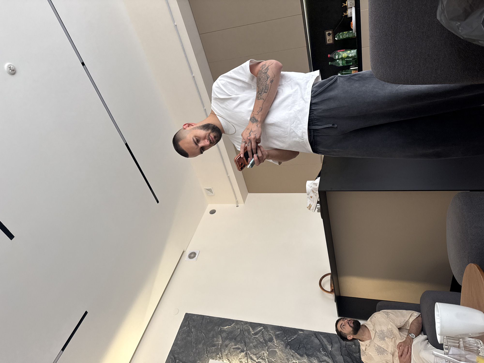 This image depicts an indoor scene, likely a modern apartment or lounge area, in Yerevan, Armenia. The space features light-colored walls, a white ceiling with linear lighting fixtures, and a smoke detector.
In the foreground, a man with a shaved head and beard stands, looking towards the camera. He is wearing a plain white short-sleeved t-shirt and dark gray sweatpants. Numerous tattoos are visible on his arms, including what appears to be a snake or dragon design and an eye. He holds a smartphone in one hand and possibly a vape pen in the other.
Further back in the scene, another man is seated on a dark gray armchair. He has a beard and is wearing a light-colored, patterned short-sleeved shirt and a watch on his left wrist. He is looking upwards, appearing relaxed or contemplative.
The background includes a dark-colored counter or cabinet unit with shelves holding several bottles, one of which displays partially visible text like "LO" and "TE," possibly indicating "LOTTE." A dark, textured art piece adorns the wall near the seated man, and a small wooden side table holds a white mug and a glass. The overall atmosphere suggests a casual, relaxed gathering, likely during the daytime.