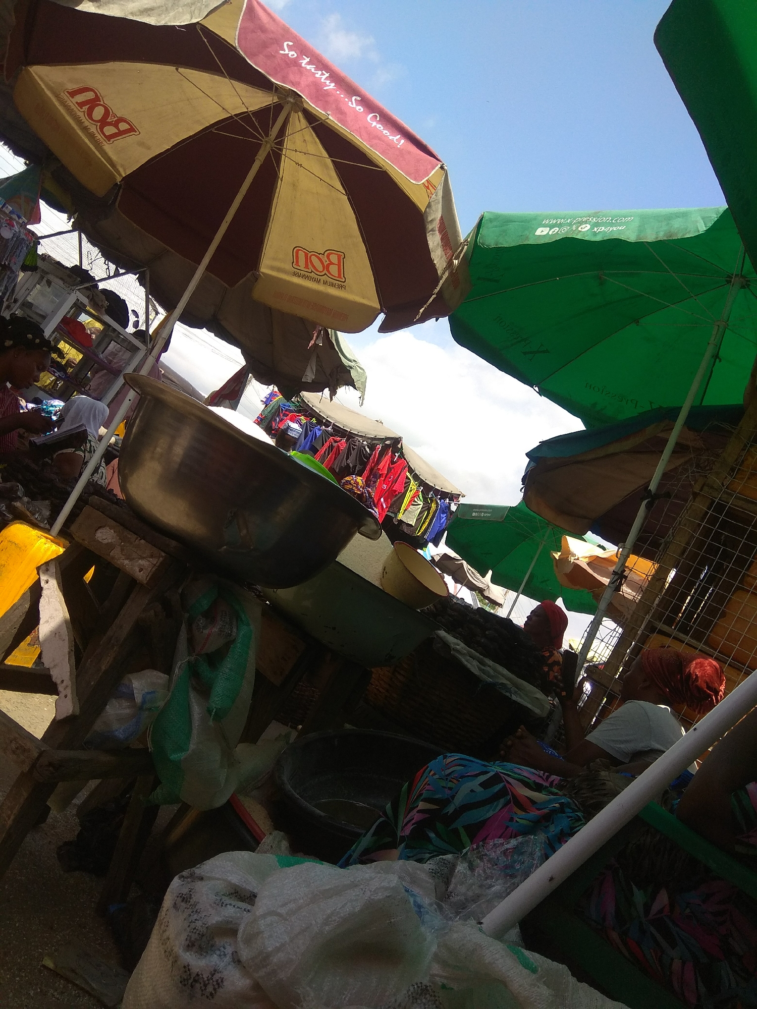 The image captures a bustling outdoor market scene in Jalingo, Nigeria, under a bright, partially cloudy sky. Several large market umbrellas are open, providing shade. A prominent maroon and yellow umbrella in the foreground displays the "Bou" logo and text that reads "So tasty... So Good!". To its right, a green umbrella features a website address and social media icons.
Beneath the umbrellas, the market is filled with various goods. In the mid-ground, a large metal basin rests on wooden supports. To the left of the basin, a woman with dark hair, wearing a striped shirt, is visible. Further left, a yellow plastic container is seen. Behind the basin, colorful clothing, including items with blue, red, and yellow hues, hangs on display.
In the foreground and mid-ground, several people are present. To the right of the metal basin, a woman with a red headscarf is partially visible, wearing a light-colored t-shirt and a vibrantly patterned skirt. Another person, also with a red headscarf, is seated on the right, facing away from the camera. Bags, some made of white and green plastic, are scattered throughout the scene, some appearing to contain goods. A white pole runs vertically in the right foreground. The ground is uneven and appears to be a mix of dirt and debris. The overall impression is one of active commerce in an open-air setting.