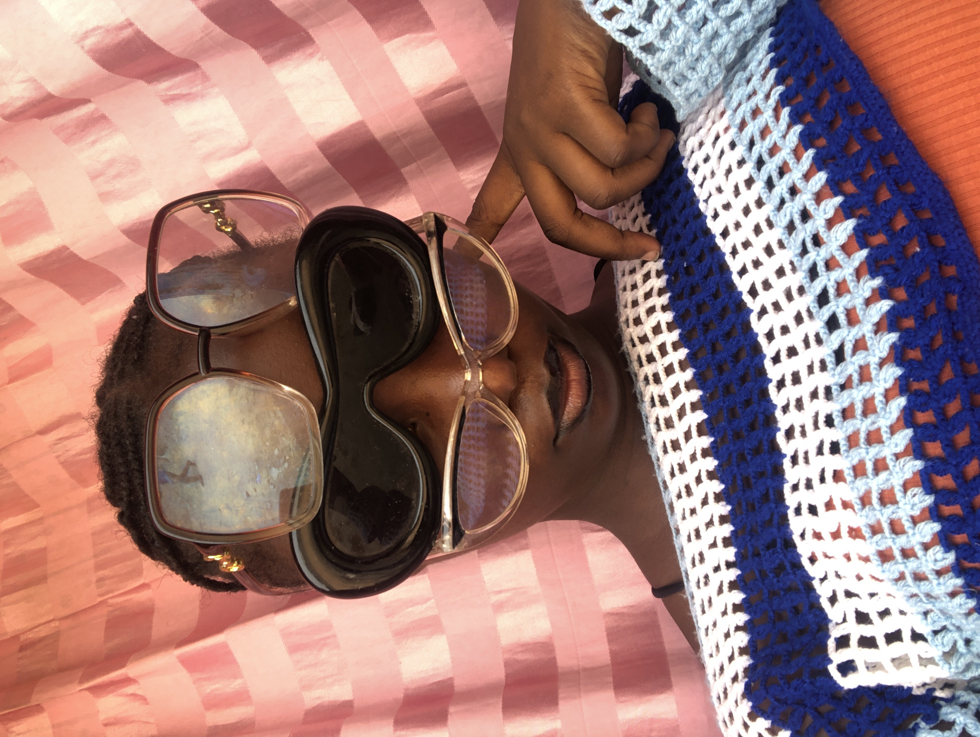 A medium shot depicts a person of African descent lying on a horizontally striped pink-and-white surface, possibly a bed or sheet. The person's head is oriented towards the left side of the frame, while their body extends out towards the right. They are wearing three pairs of sunglasses: two pairs with large, square frames, one in gold and the other with clear frames, and a third pair with black, asymmetrical lenses.
A hand is visible near the person's head, suggesting they are adjusting or interacting with the sunglasses. They are wearing a crocheted garment featuring a pattern of white, light blue, and dark blue stripes, which covers part of their body on the right side of the frame.
The background appears to be a simple indoor setting. The image was likely taken in Akwanga, Nigeria.