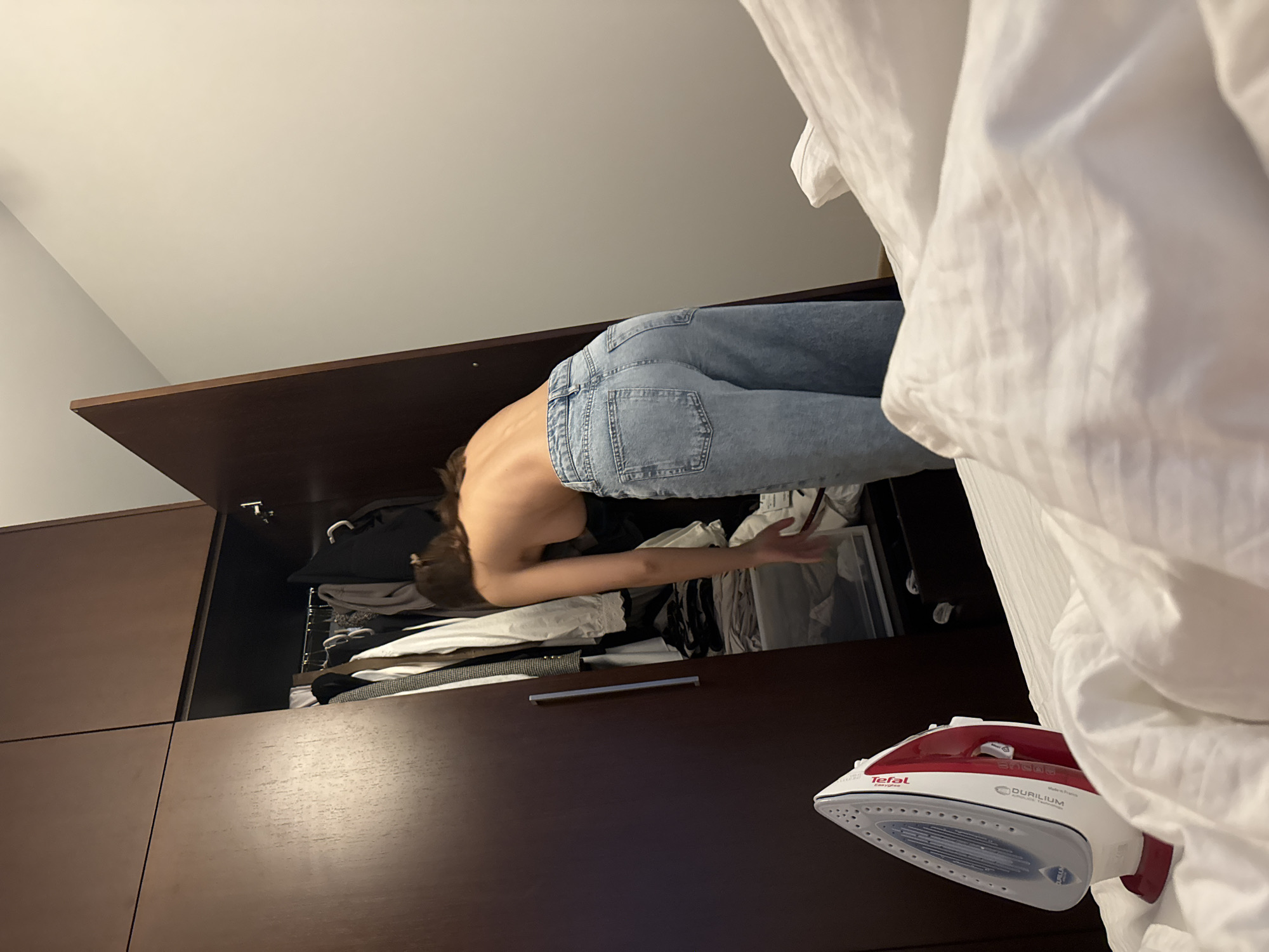 An indoor scene depicts a person reaching into a partially open dark wood wardrobe, located in the city of Unknown, Unknown. In the immediate foreground, a red and white Tefal steam iron stands upright on white textured bedding. A female individual, seen from a slightly elevated angle from behind and the right side, is positioned facing the open wardrobe. She is shirtless, exposing her back, right shoulder, and side. She wears light blue, loose-fitting denim jeans. Her brown hair is pulled back. Her right arm extends into the lower section of the wardrobe, suggesting she is retrieving an item. The wardrobe interior reveals various clothes hanging on hangers and folded items in clear storage containers at the bottom. To the right of the person, a plain light-colored wall is visible.