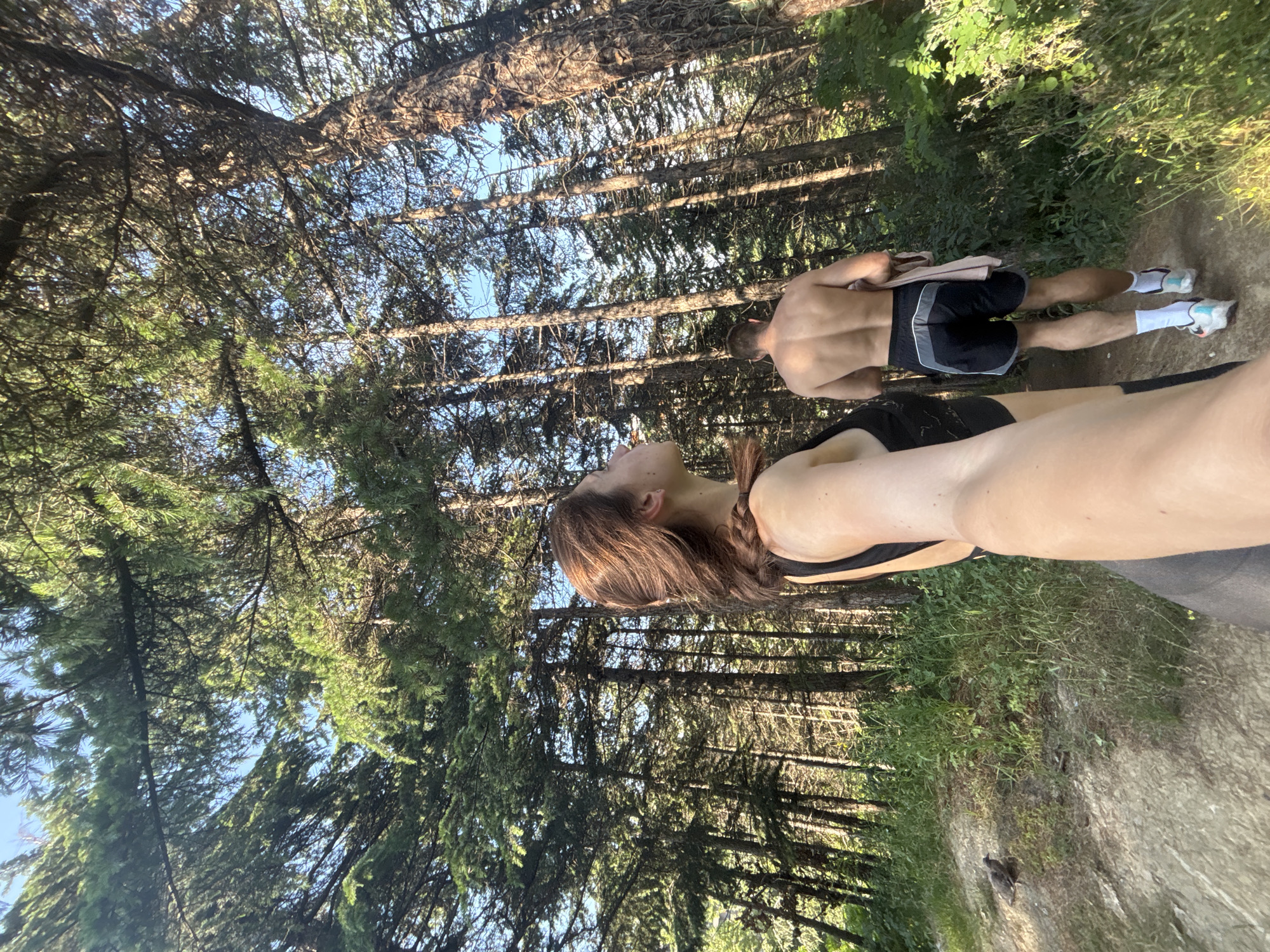 The image depicts a selfie taken by a woman from the chest up, with her arm extended to capture herself and a man walking away from the camera. They are in a dense forest with tall, slender pine trees. The woman has brown hair, pulled back, and is wearing a black sports bra. The man is shirtless, revealing his muscular back, and is wearing black athletic shorts with grey and white trim, white socks, and athletic shoes. He is holding a light pink towel over his right shoulder. The path they are walking on appears to be dirt and gravel, with green grass and some yellow wildflowers growing on the sides. The sun is shining through the trees, creating dappled light. The sky is visible between the branches and is a clear blue. The location is not specified as Varna, Bulgaria, and there are no identifiable landmarks to suggest this location.