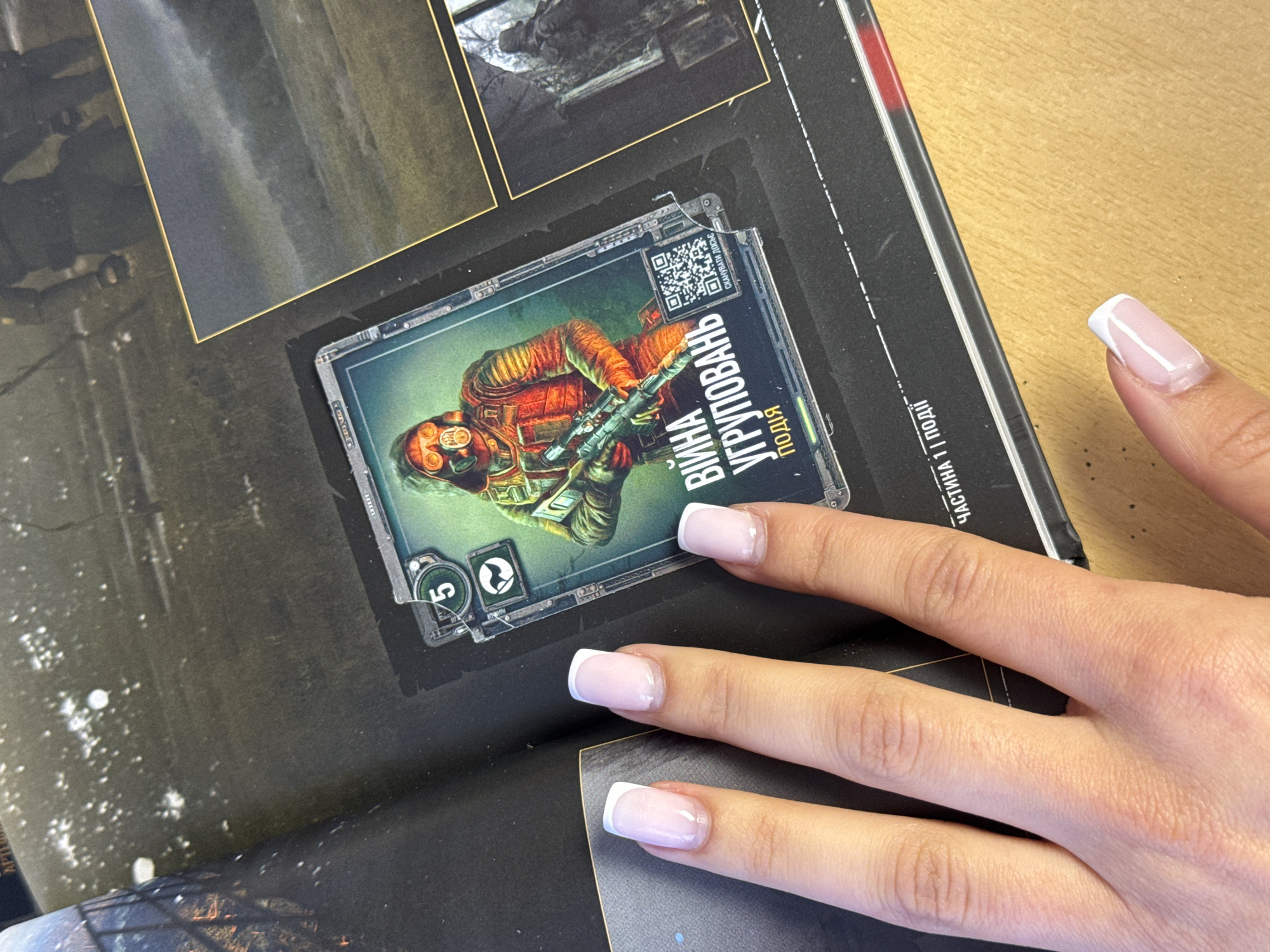 A human hand with a French manicure rests on a book that is open to a page with a printed image. The image appears to be a trading card or an illustration from a game, featuring a figure in protective gear, holding a weapon, with Ukrainian text and a QR code. The book appears to be on a wooden table. The location cannot be determined from the image.