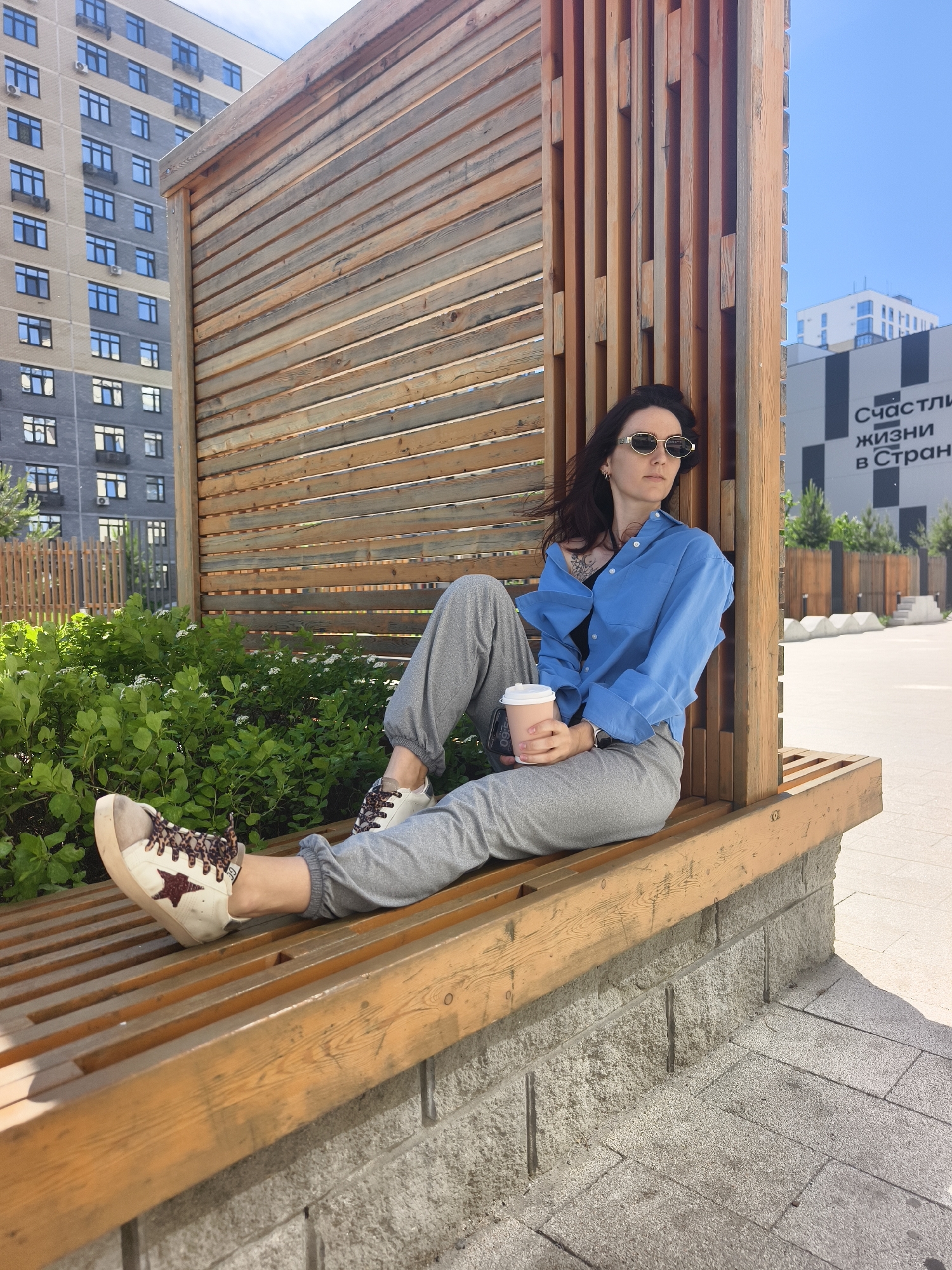 A young woman is seated on a wooden slatted bench within an urban outdoor setting. She has dark hair, wears sunglasses, a light blue long-sleeved shirt over a dark top, and grey sweatpants. Her left foot, clad in a white and beige sneaker with a dark star detail and patterned laces, rests on the bench. She holds a light-colored disposable coffee cup in her right hand and is looking towards the right side of the frame. A small tattoo is visible on her left shoulder.
The bench is part of a larger wooden structure, possibly a pergola or decorative wall, featuring horizontal and vertical wooden slats. Behind her to the left, there are lush green bushes with small white flowers. In the background, modern multi-story buildings are visible under a clear blue sky. One building to the far left is light brown and grey with numerous windows. Another building to the right, partially obscured, has black and white geometric patterns and features the Russian text "Счастливой жизни в Стране" (Happy life in the Country/Land). The ground around the bench is paved with light grey tiles. The scene appears to be taking place on a bright, sunny day, indicating good weather. The overall setting suggests a contemporary public space or residential area.