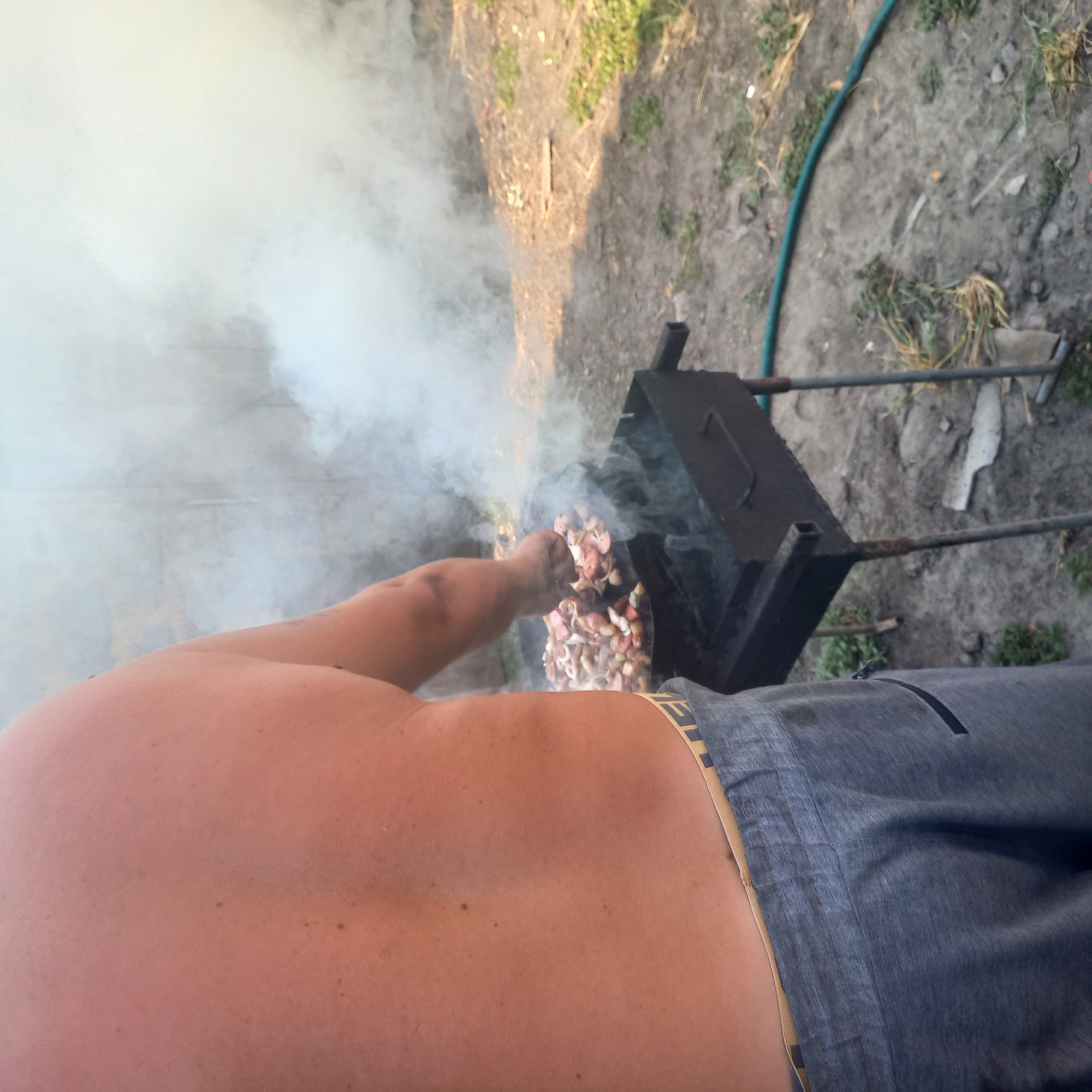 An individual, shirtless and wearing grey sweatpants, is cooking meat on a black charcoal grill in an outdoor setting. The grill contains a pan filled with chopped meat, which is emitting a significant amount of smoke. The ground around the grill appears to be dirt and is sparsely covered with dry grass and some green vegetation. A green garden hose is visible on the ground. The background is a blurred, light-colored wall or structure. The lighting suggests it is daytime. The image quality is moderate, with some graininess. The exact location within the city of Klyntsi, Ukraine, cannot be determined from the visual information provided.