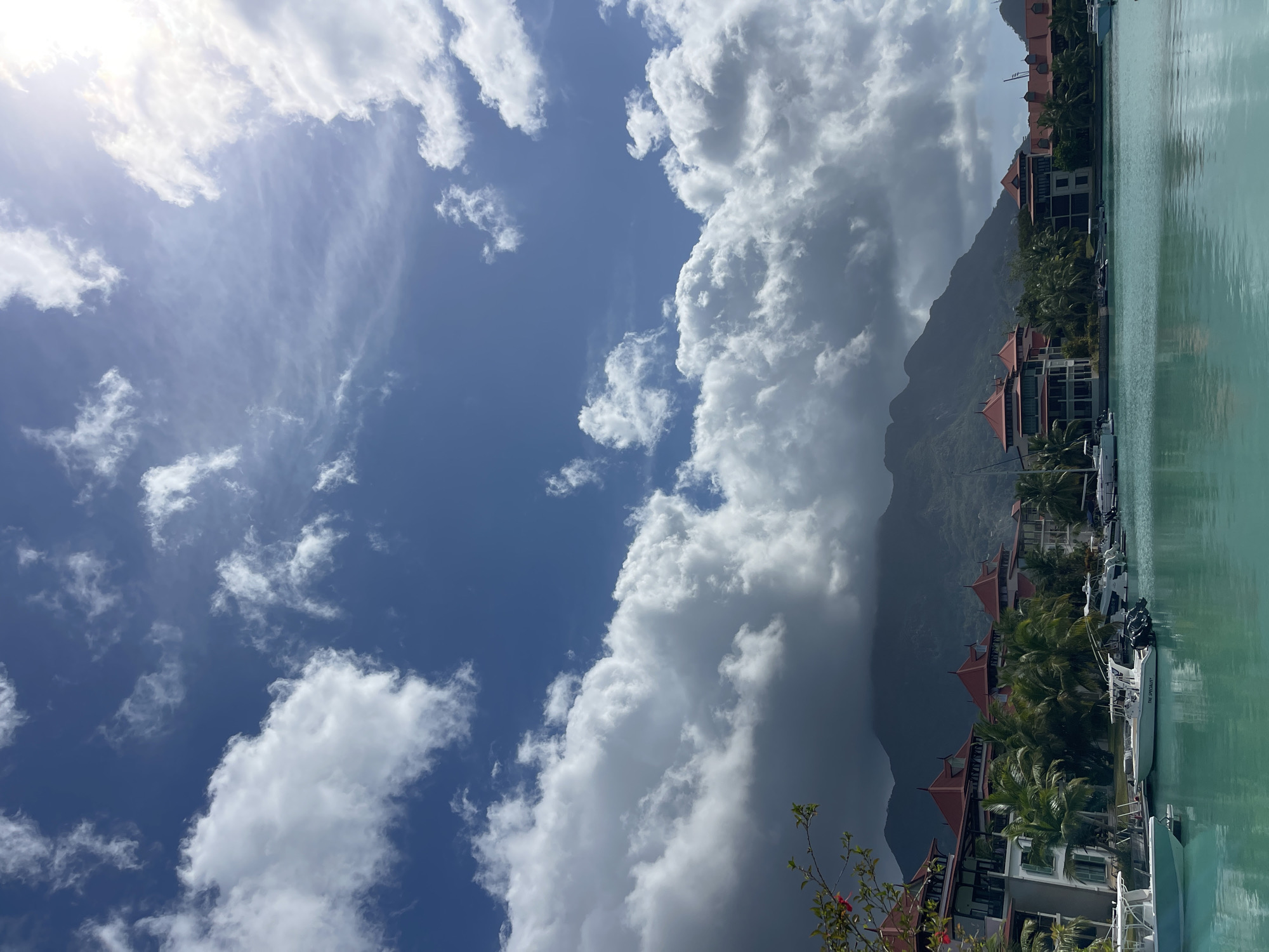 The image depicts a waterfront scene in Providence, Seychelles, featuring a marina with several boats docked. In the background, there are multi-story buildings with distinctive red-tiled roofs, characteristic of tropical architecture. These buildings are surrounded by lush palm trees. Beyond the buildings, a mountain range dominates the horizon, partially obscured by dramatic, voluminous clouds. The sky is a vibrant blue, with a mix of white, puffy clouds and wispy cirrus formations, suggesting a partly cloudy day with bright sunlight. The water in the foreground is a clear, turquoise hue, reflecting the sky and the surrounding environment. The scene conveys a sense of tropical luxury and natural beauty.