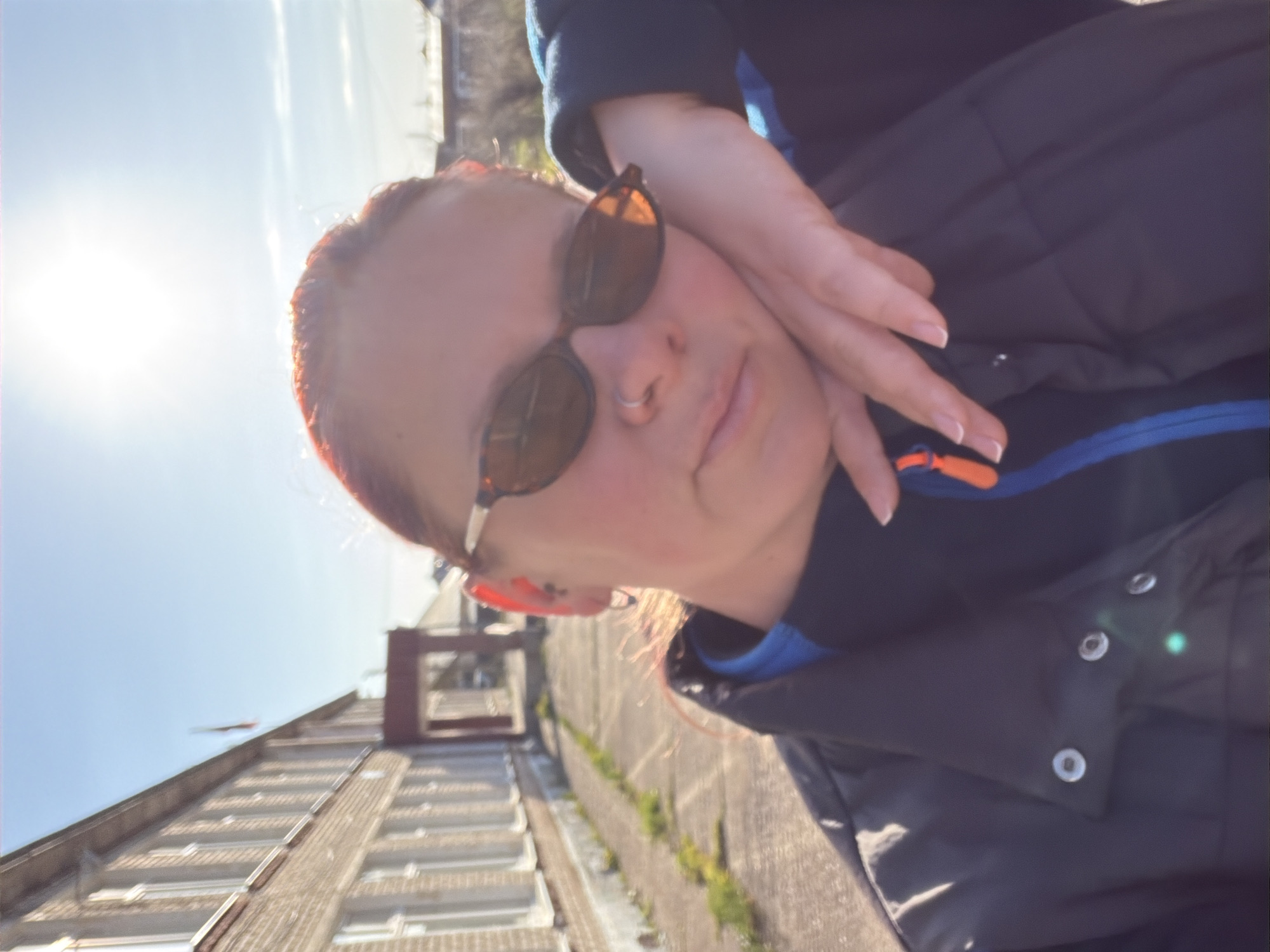 A medium shot photograph captures a woman in Chaikovsky, Russia. She wears sunglasses and a dark-colored jacket with blue trim and an orange zipper pull. Her hand is raised near her face. Behind her, a brick building with a window grid is visible, along with a clear blue sky and bright sunlight. A low concrete wall or fence with some vegetation is also in the background.