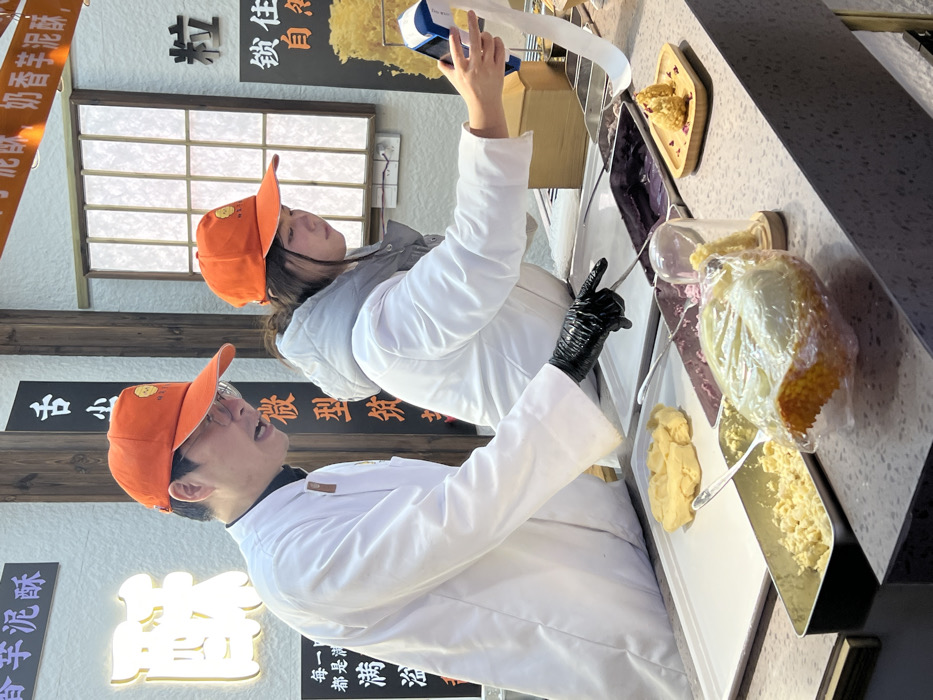 An indoor scene in Yuzhong District, China, depicts two individuals preparing food at a counter. On the left, a male wearing an orange baseball cap, glasses, and a white chef's coat gestures with his gloved left hand towards a rectangular metal tray containing a purple paste. His mouth is open, suggesting speech. On the right, a female, also in an orange baseball cap and a white hooded jacket, operates a white and blue portable device that dispenses a long strip of paper. The gray speckled countertop displays various food items and ingredients: a lump of yellow dough on a white cutting board, another rectangular metal tray with a yellowish granular substance, and a large durian-shaped dessert wrapped in clear plastic. A small wooden plate holds two golden-brown, conical pastries garnished with pink petals. The background features light-colored walls with illuminated Chinese signage and a traditional-style window with translucent panels.