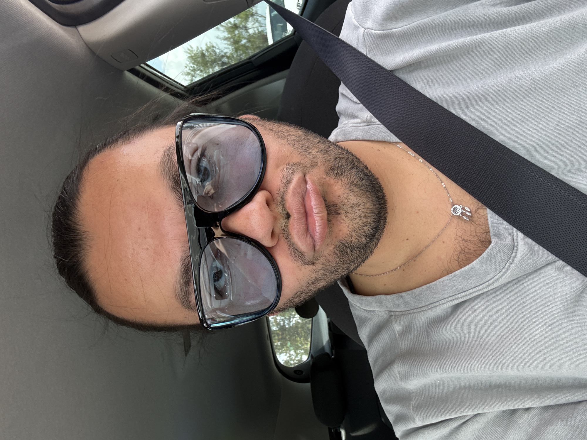A person wearing glasses and a gray shirt sits in a car with a seatbelt fastened. The car has a sunroof. The location is undetermined.