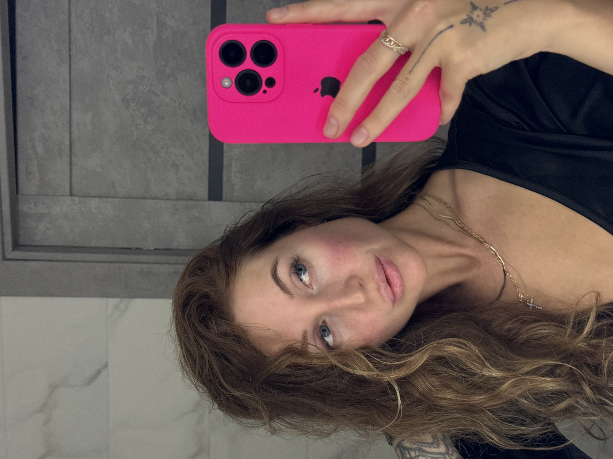A woman with light brown, curly hair and blue eyes is taking a selfie. She is wearing a black top and a gold necklace with a cross pendant. She is holding a pink iPhone with a triple-lens camera in her right hand. The back of her hand has a tattoo. She is wearing a ring on her finger. The background consists of gray and white wall tiles. There is no evidence in the photograph to suggest the location is in Chaikovsky, Russia.