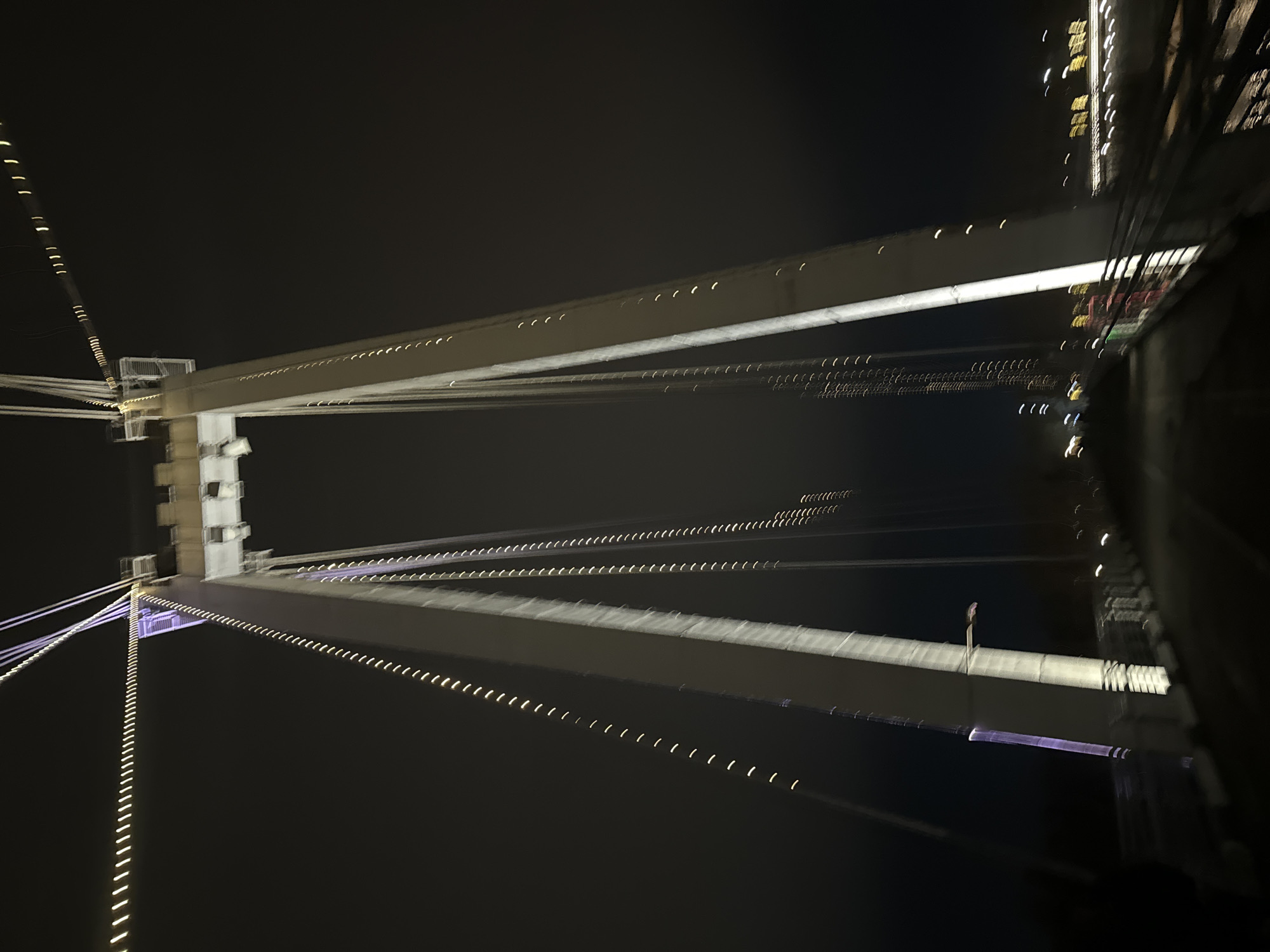The image depicts an upward view of a bridge structure at night. The bridge features prominent white beams, likely suspension or support elements, adorned with evenly spaced lights. The background is a dark, mostly featureless sky. The angle suggests the picture was taken from underneath the bridge, looking up at the structure and its lighting. The scene could possibly be Komunalny Bridge in Krasnoyarsk, Russia. The scene appears blurred due to motion or a long exposure time.