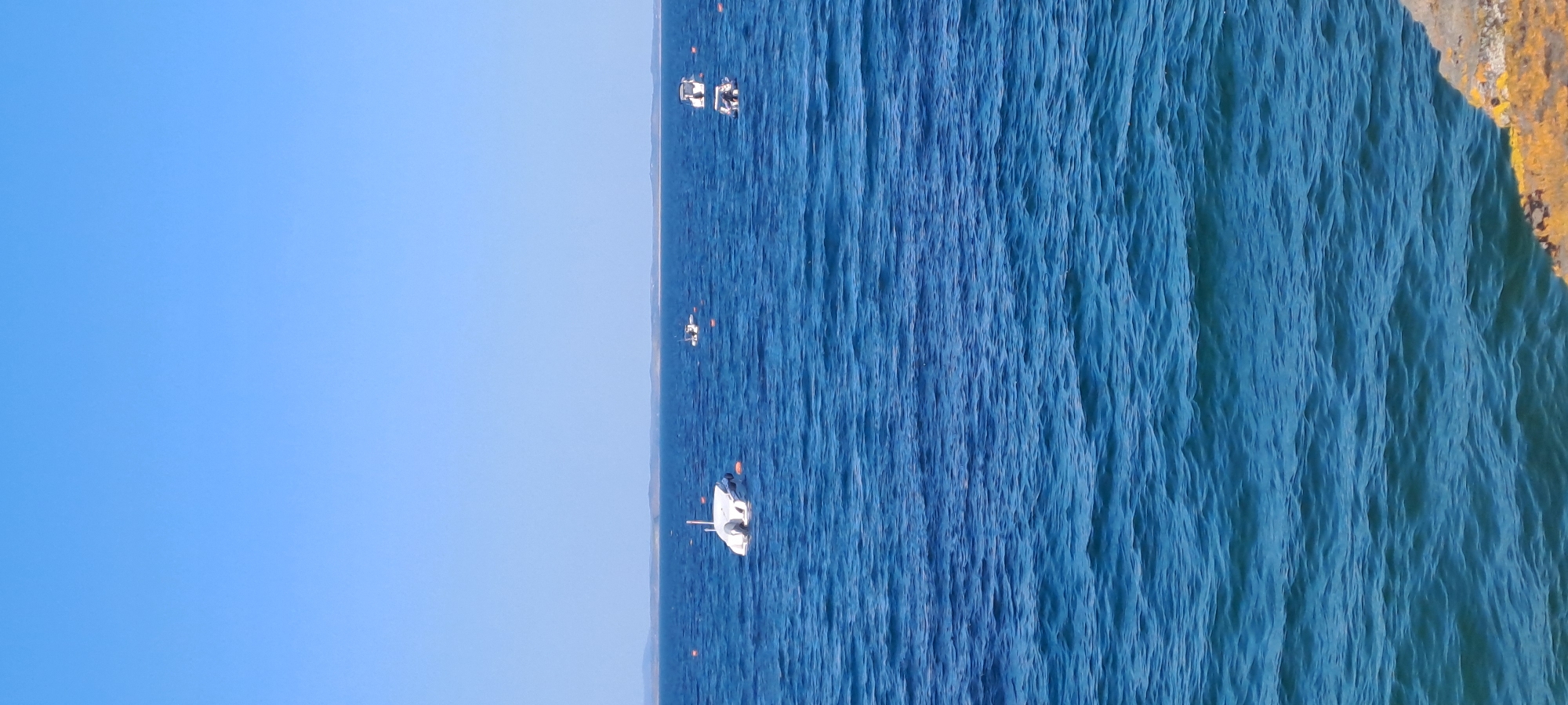 The image shows a calm blue sea with a clear sky. Several boats are visible on the water. In the foreground, there is a rocky shore. In the distance, a faint landmass can be seen. The scene appears to be a serene coastal view. The location is unknown, Ireland.