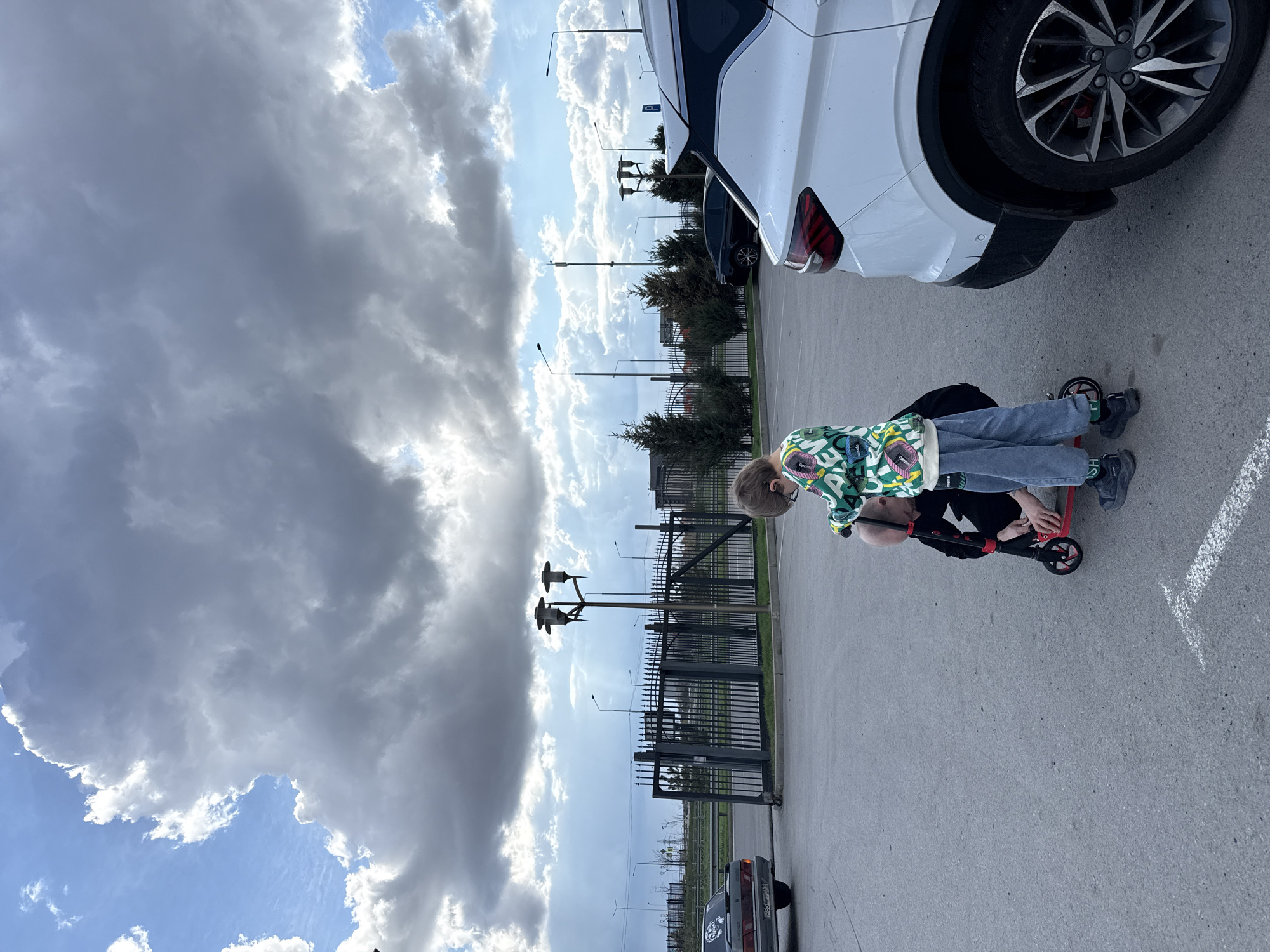 Two children are on a red scooter on an asphalt surface. One child wears a green patterned jacket and blue jeans. The other child wears a black top. A white SUV is parked nearby. A metal fence borders the paved area. Utility poles and street lights are visible in the background. A grey car is parked in the distance. A cloudy sky is overhead. The location is Rostov-on-Don, Russia.