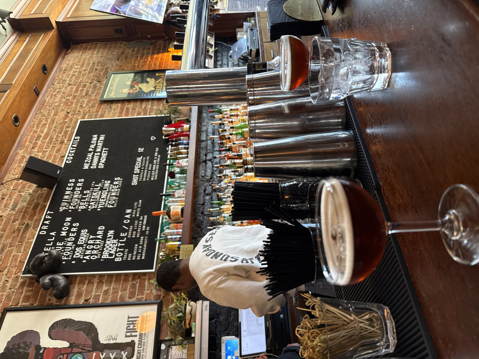 The image depicts the interior of a bar in New York City, United States of America.
The scene focuses on a wooden bar counter and the area behind it. A male individual, appearing to be a bartender, is positioned behind the bar, facing away from the viewer. He wears a light-colored sweatshirt with "SUNDAY SERVICE" printed in an arc across the back. He is looking down at a computer screen to his left.
Behind the bartender, a black brick wall forms the base, topped by a wooden shelf holding numerous liquor bottles of various shapes and colors. Above this, an exposed red brick wall extends upwards. Mounted on the brick wall is a large black chalkboard menu displaying "DRAFT," "BOTTLE / CAN," and "COCKTAILS" sections with various drink options, including "Mezcal Paloma," "Danny's Martini," and "Spaghetti." A "Shot Special 12" is also listed. To the left of the menu, a framed poster depicting a stylized, cactus-like figure with "FIGHT CHAMPIONSHIP" visible is hanging, along with a pair of black boxing gloves. To the right, another framed artwork partially obscured with "Now Available!" is visible. Recessed lighting is present in the wooden ceiling. A television screen displaying colorful content is partially visible on the far right.
On the wooden bar counter in the foreground, two cocktails are prominently featured. Closest to the viewer, a coupe glass holds a dark amber liquid topped with a thin layer of foam. To its right, an elevated, wider coupe glass contains a similar dark amber liquid. Adjacent to this, a short, faceted glass filled with ice cubes and melted water is present. Several stainless steel cocktail shakers of varying sizes are arranged on a black mat behind these drinks. A container full of black drinking straws and a clear glass vase-like container filled with numerous bamboo cocktail skewers with looped ends are also visible on the bar.